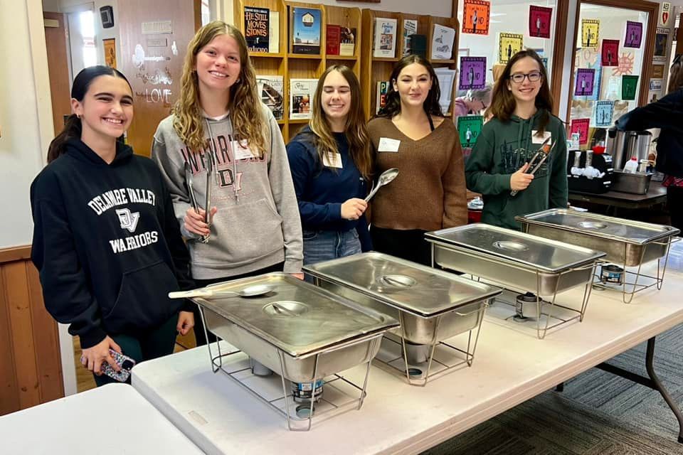 Five young people stand behind a table with food warmers. They smile, holding serving utensils, in a room with books.