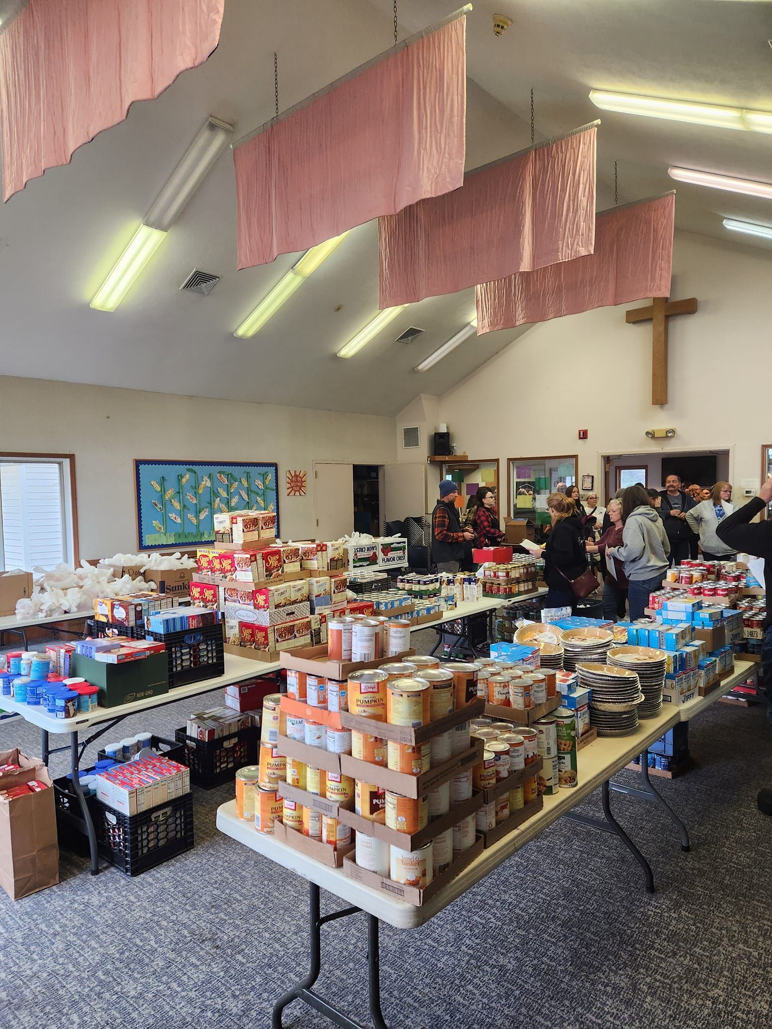Food pantry with tables full of canned goods, volunteers, and people. A cross hangs on the wall.