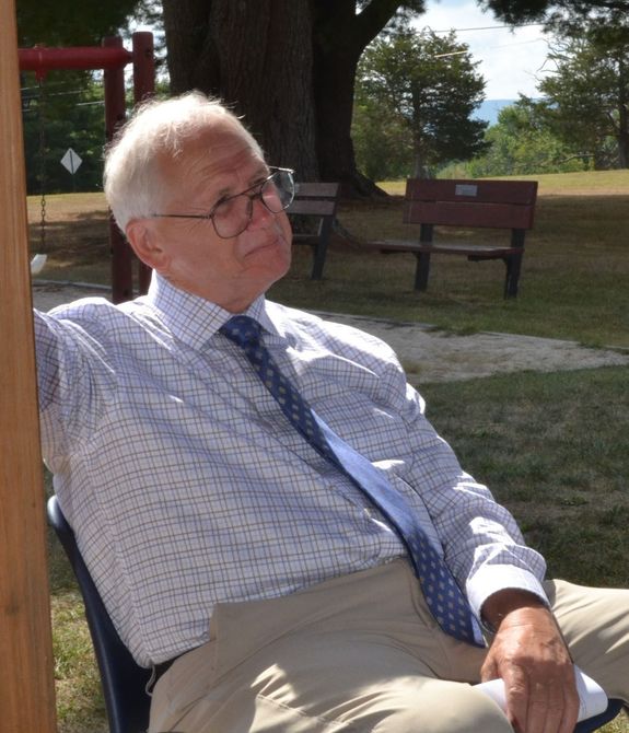 Older person wearing glasses, blue tie, and collared shirt sits outdoors, looking off to the side, near benches and trees.