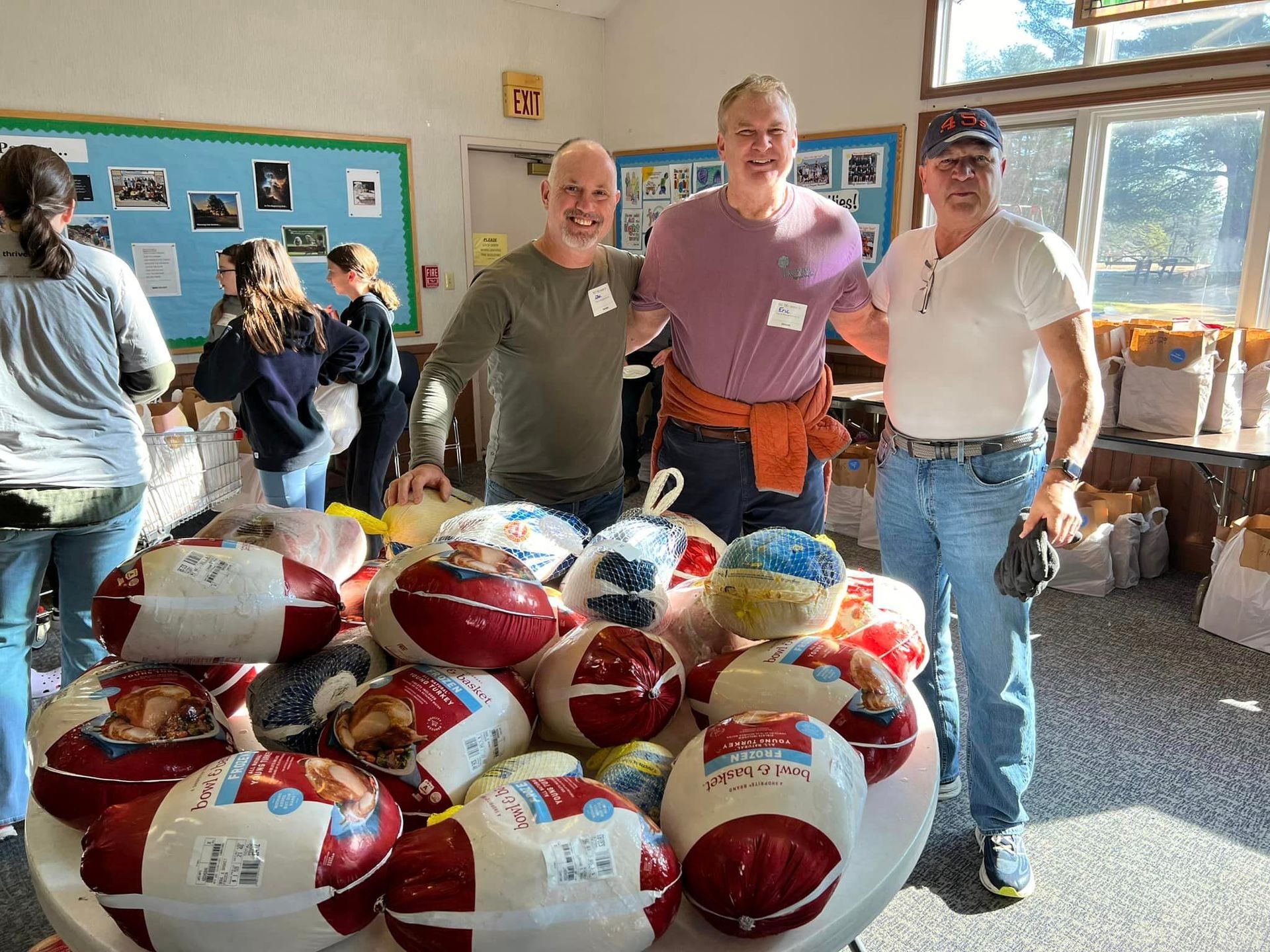Three men pose with a pile of frozen turkeys in a room. People in background.