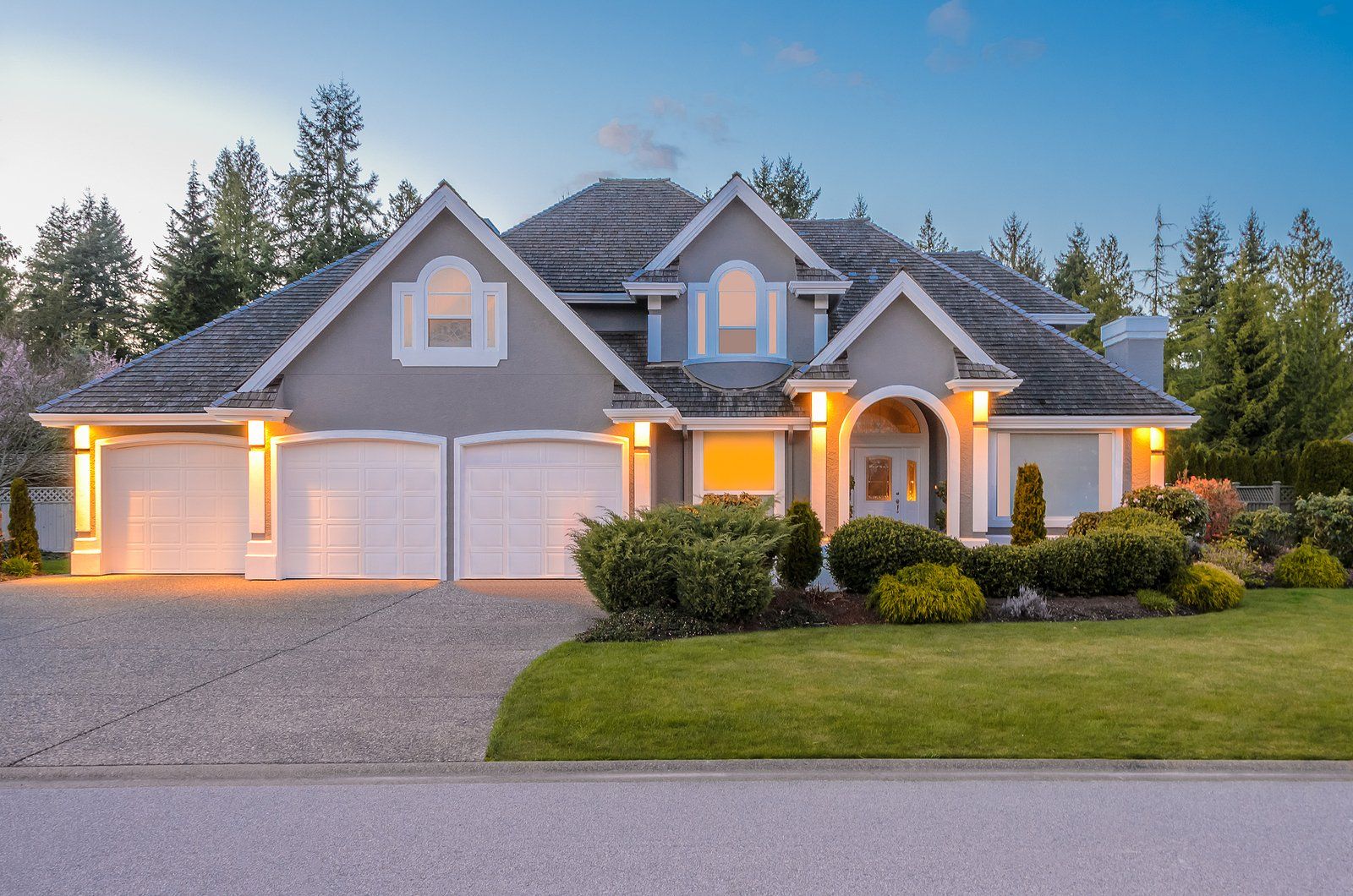 Large gray suburban house with lit garages, manicured lawn, and dusk sky in the background