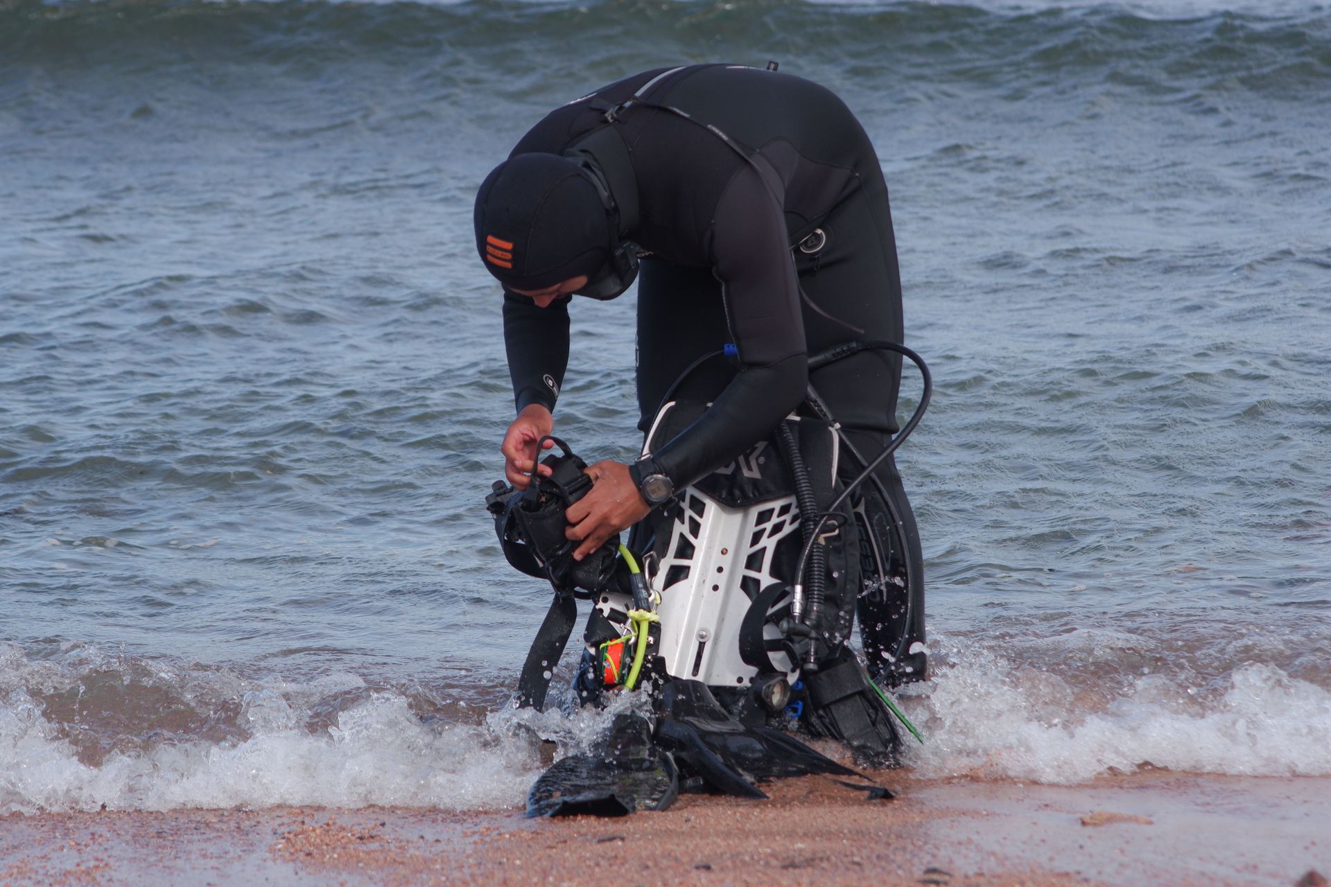 Person in a wetsuit adjusting a black-and-white prosthetic leg at the shoreline while waves wash in.