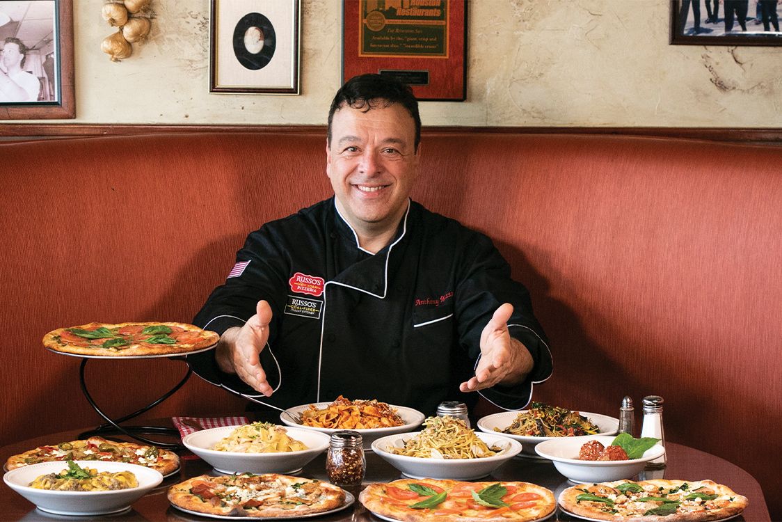 Russo's smiling, surrounded by various Italian dishes, at a restaurant table.