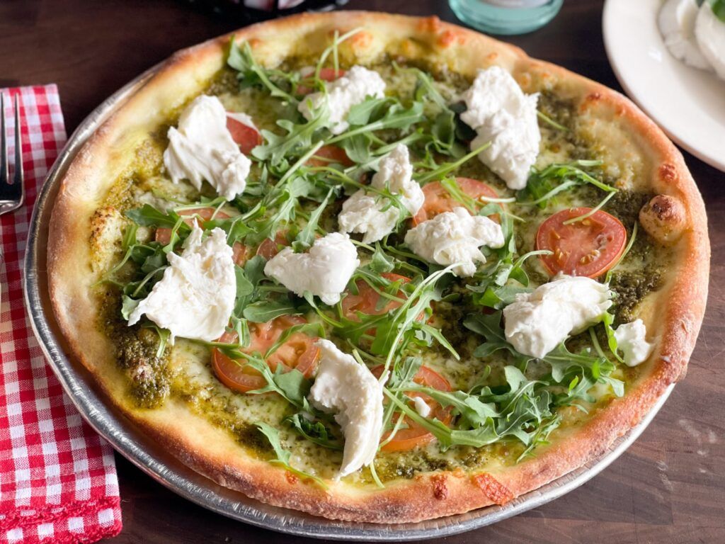 Pizza with pesto, ricotta, arugula, and tomatoes on a metal pan.
