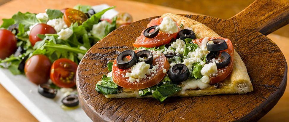 Pizza slice topped with olives, tomatoes and feta cheese on a wooden serving board, next to a salad.