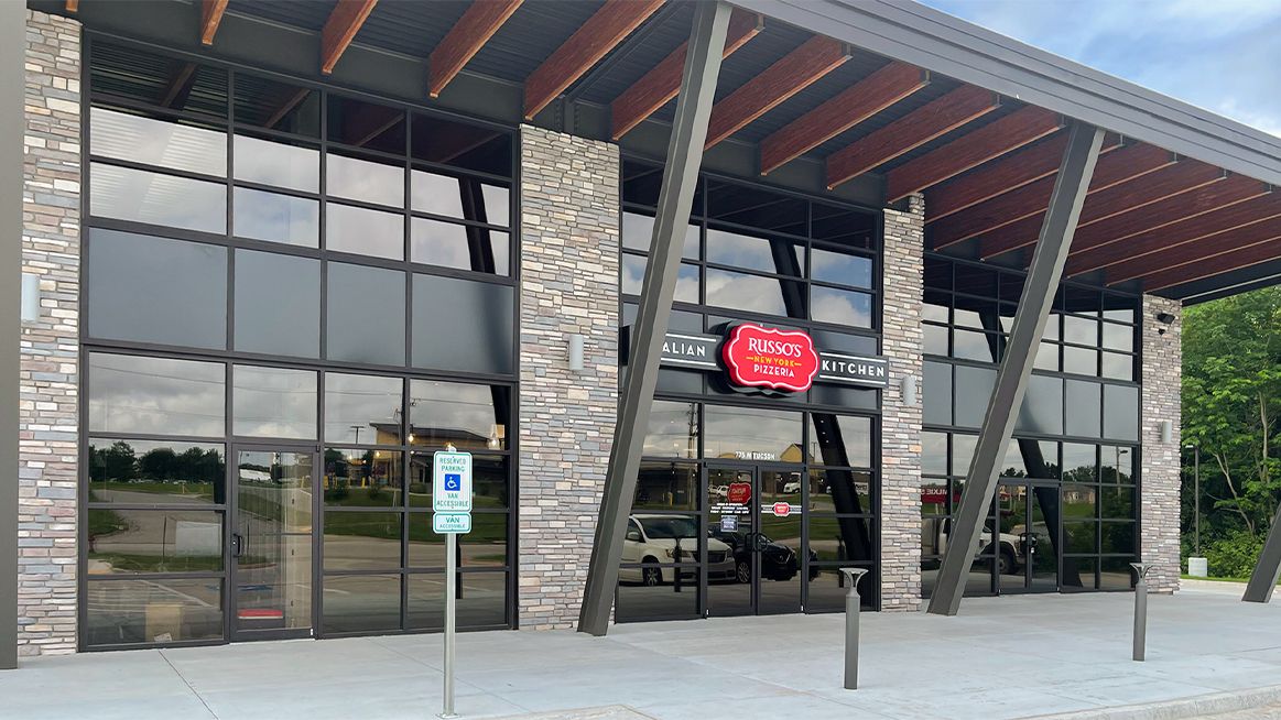 Exterior of a modern restaurant with large windows, stone accents, and a covered entrance. A sign identifies the business.