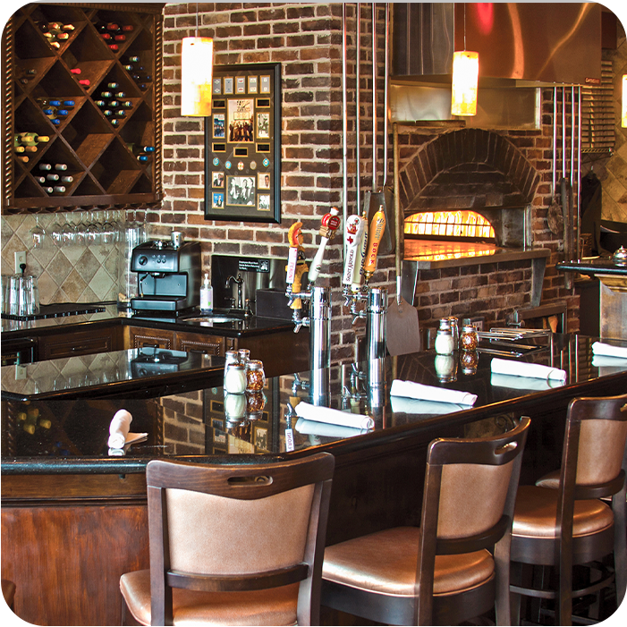 A bar with dark wood and a black countertop. Brown barstools line the front, with a pizza oven visible in the background.