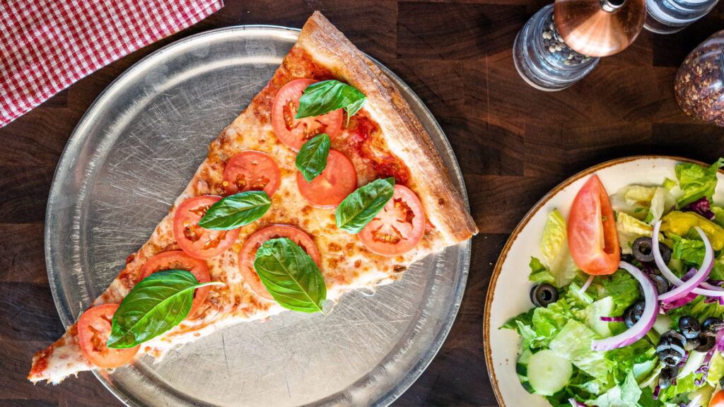 Slice of pizza with tomatoes and basil on a pizza pan, salad on the side.