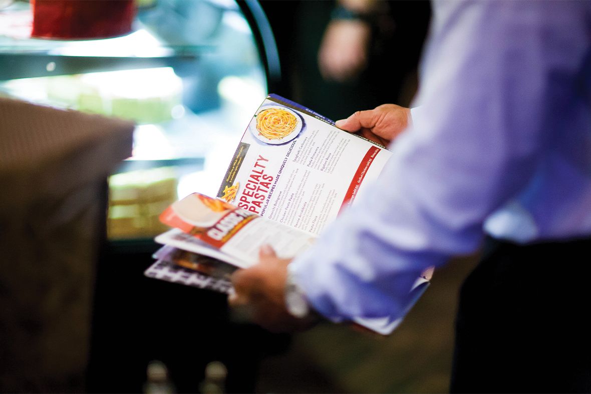 Person holding a stack of food menus near a glass display case, likely in a restaurant. The menus feature a colorful dish.