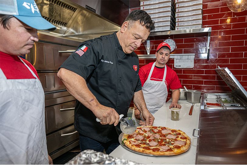 Chef in black uniform slicing pizza in a kitchen, two other workers in aprons watching.