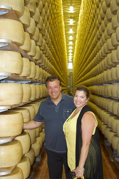 A man and woman stand between rows of aging cheese wheels in a cheese cellar. 