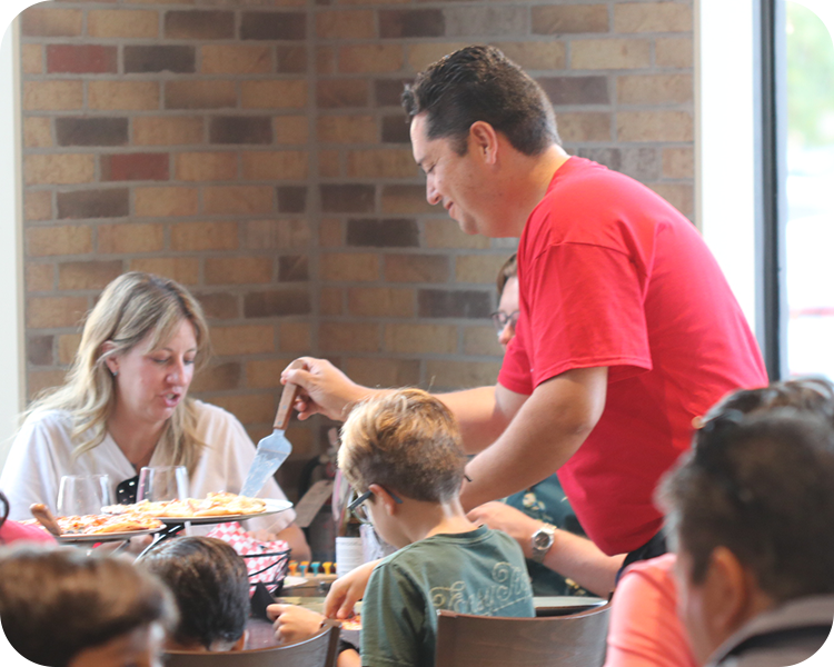 A man in red shirt serving pizza to a family at a restaurant table near a window.