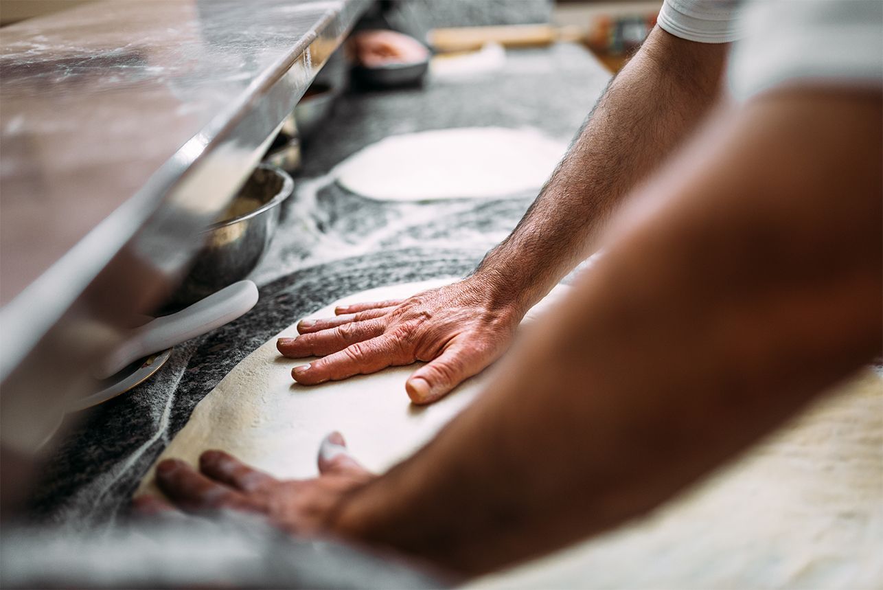 Hands of a person shaping pizza dough on a floured countertop in a commercial kitchen.
