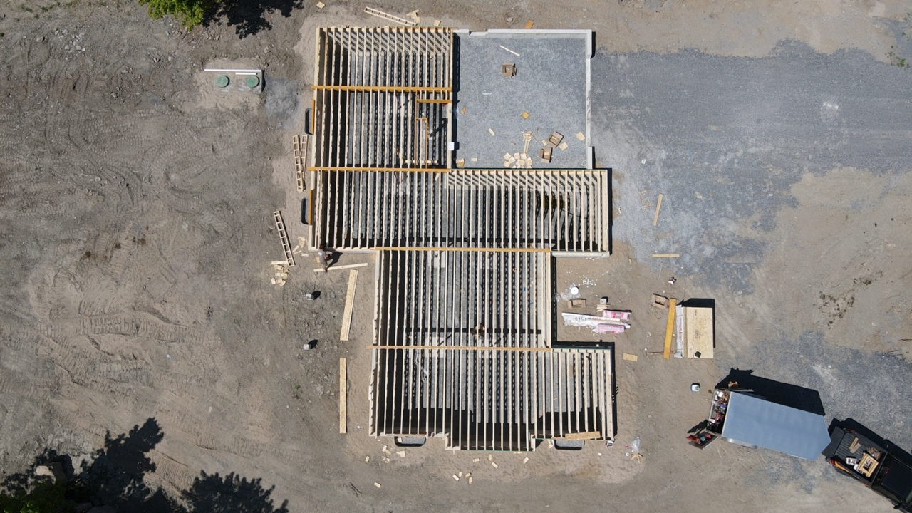 Vue aérienne d'un chantier de construction avec charpente apparente, outils et un véhicule sombre à proximité, sur un sol en terre battue.