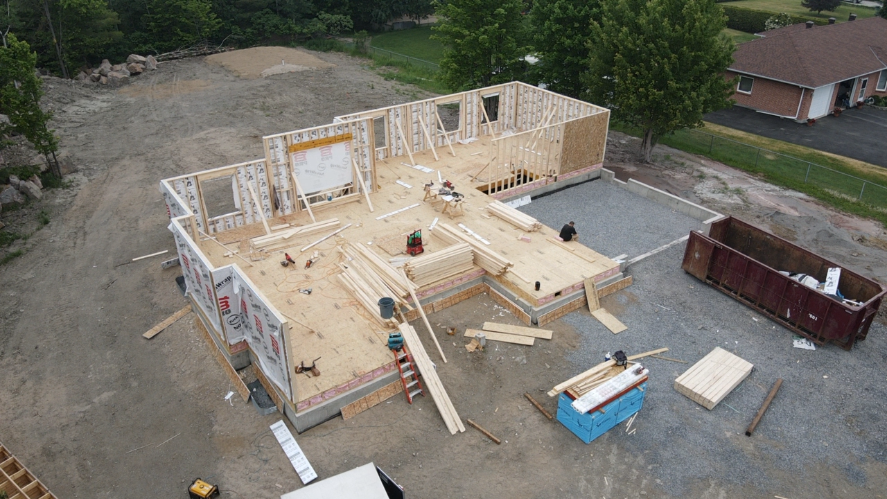 Vue aérienne d'une maison en construction avec une charpente en bois apparente, un terrain gravillonné et une benne à ordures à proximité.