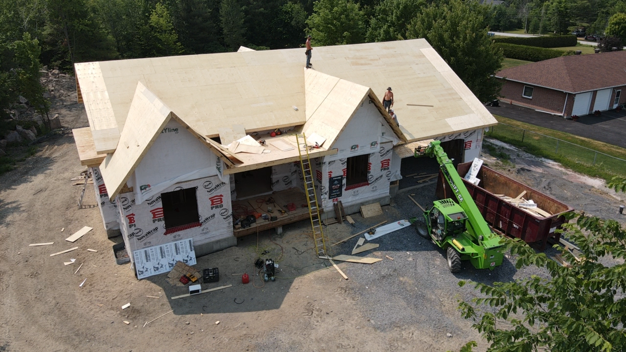 Vue aérienne d'une maison en construction, avec la charpente apparente du toit et des engins de chantier à proximité.