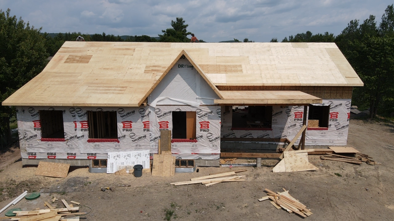 Maison partiellement construite, recouverte d'un revêtement blanc et dotée d'un toit neuf, sur un terrain en terre battue sous un ciel dégagé.