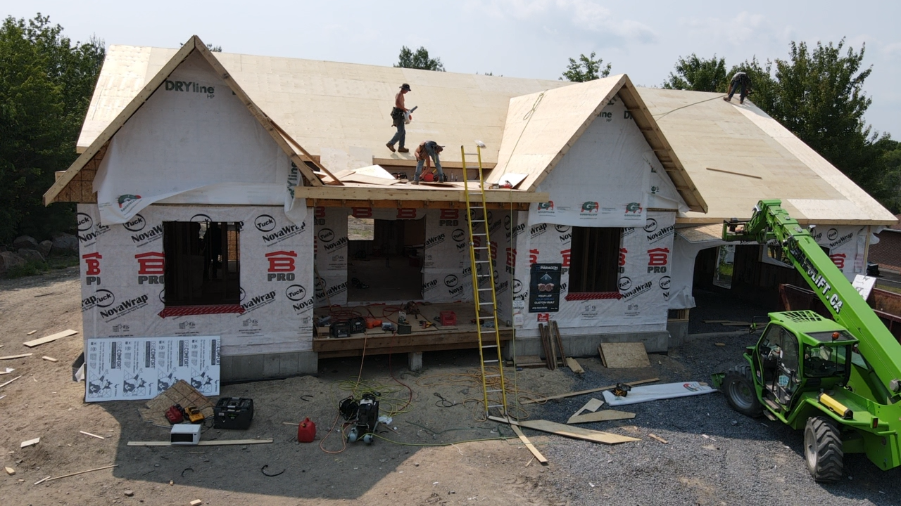 Maison en construction avec des ouvriers sur le toit et un ascenseur vert à côté.