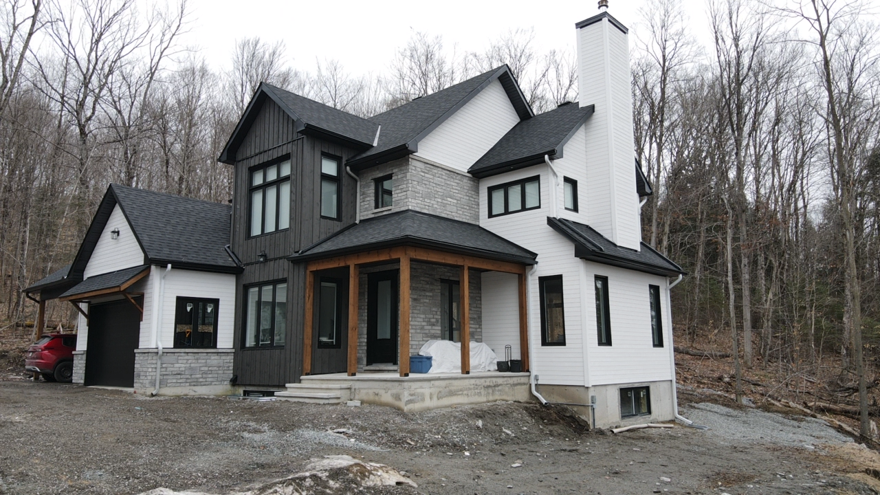 Maison moderne blanche et noire avec un porche, située sur un terrain nu à côté d'arbres dénudés.