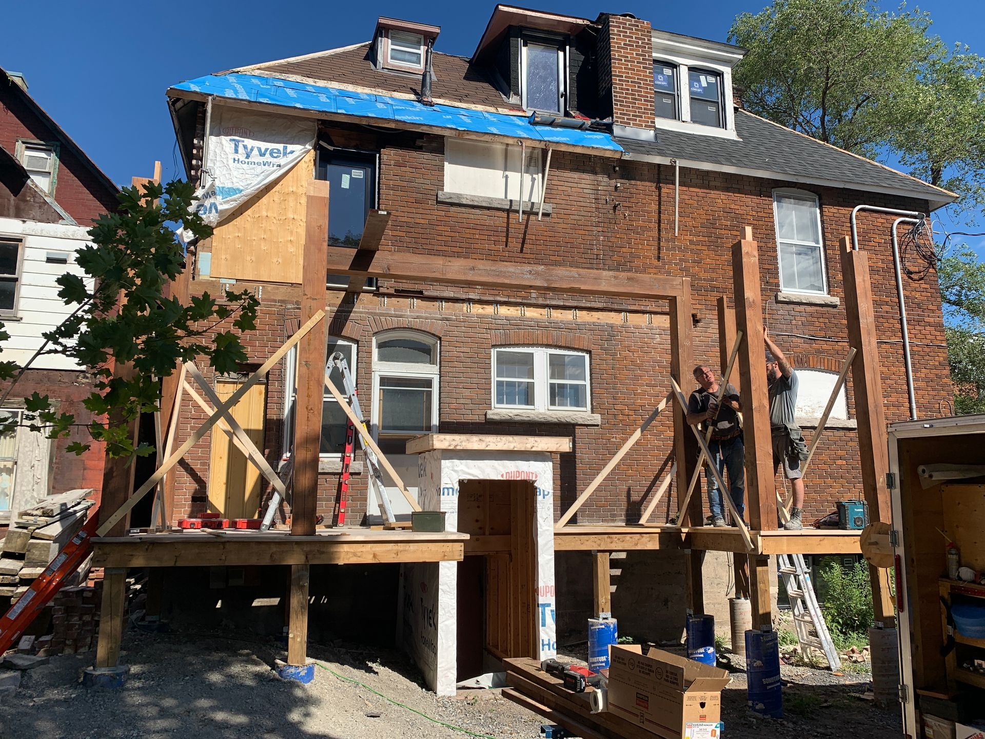 Maison à ossature bois en rénovation, avec des ouvriers, des échafaudages et un toit en bâche bleue sous un ciel dégagé.