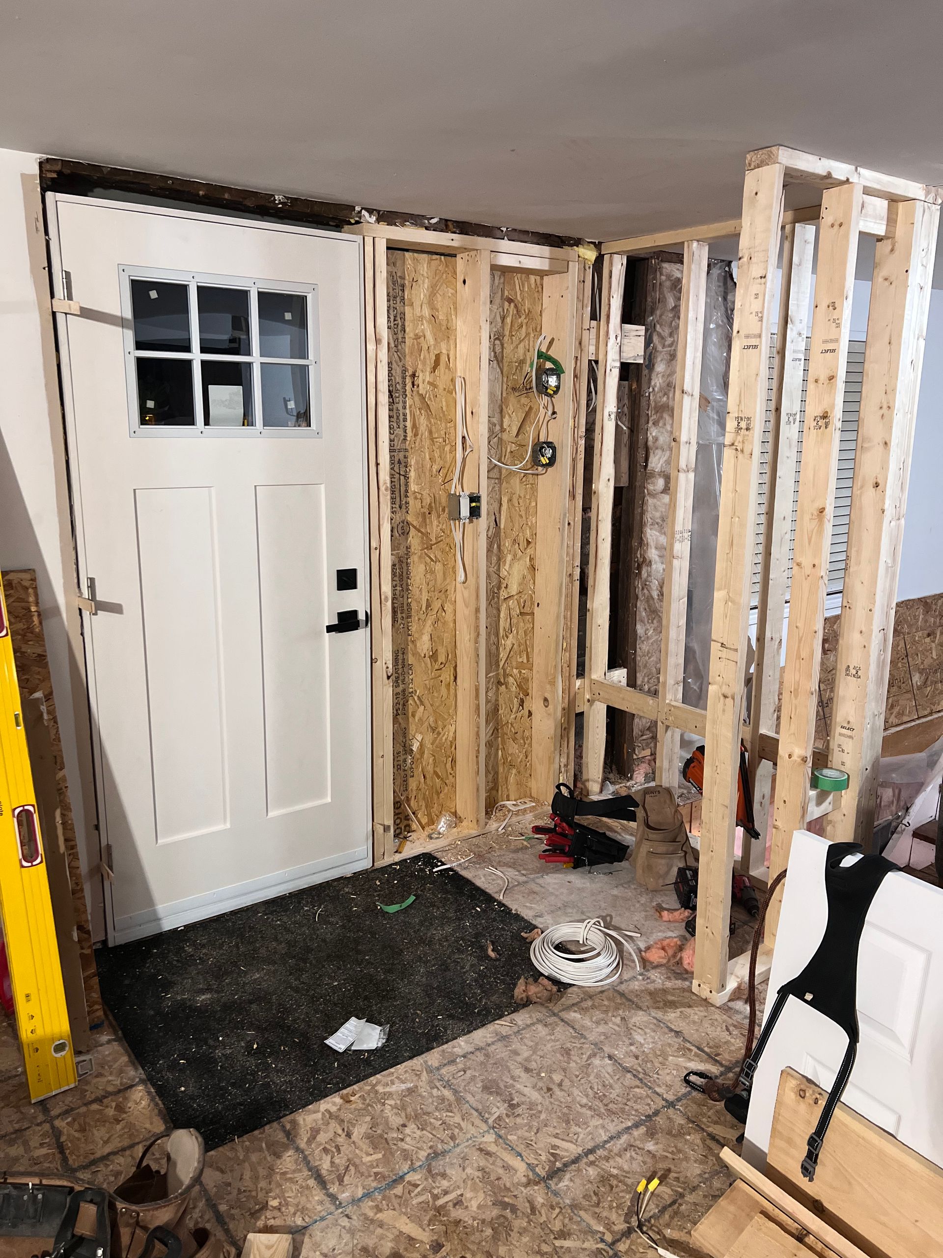 Entrée intérieure en cours de rénovation, avec montants muraux apparents et porte d'entrée blanche.