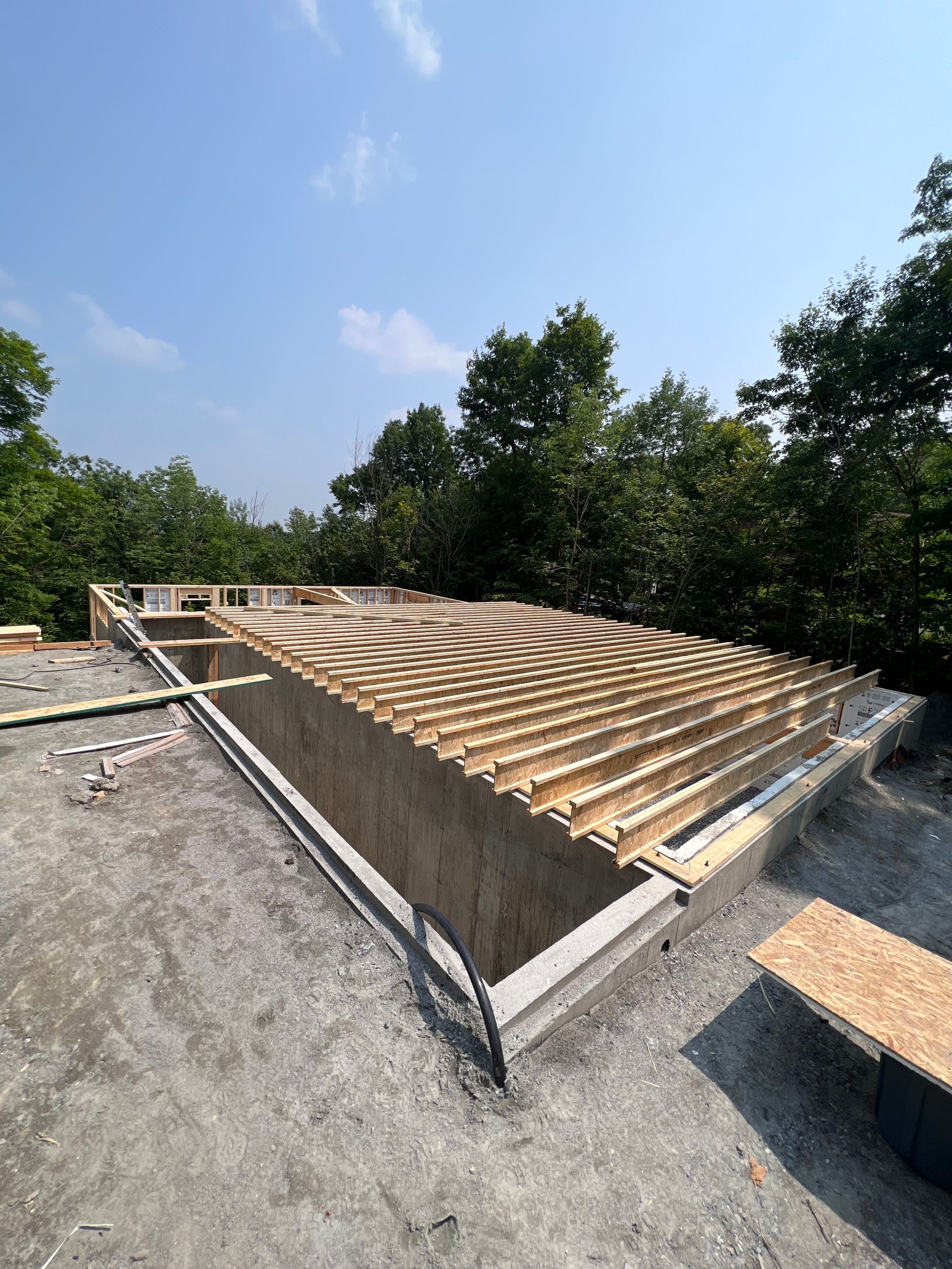 Chantier de construction avec une grande charpente de toit en bois inclinée sous un ciel bleu, entouré d'arbres