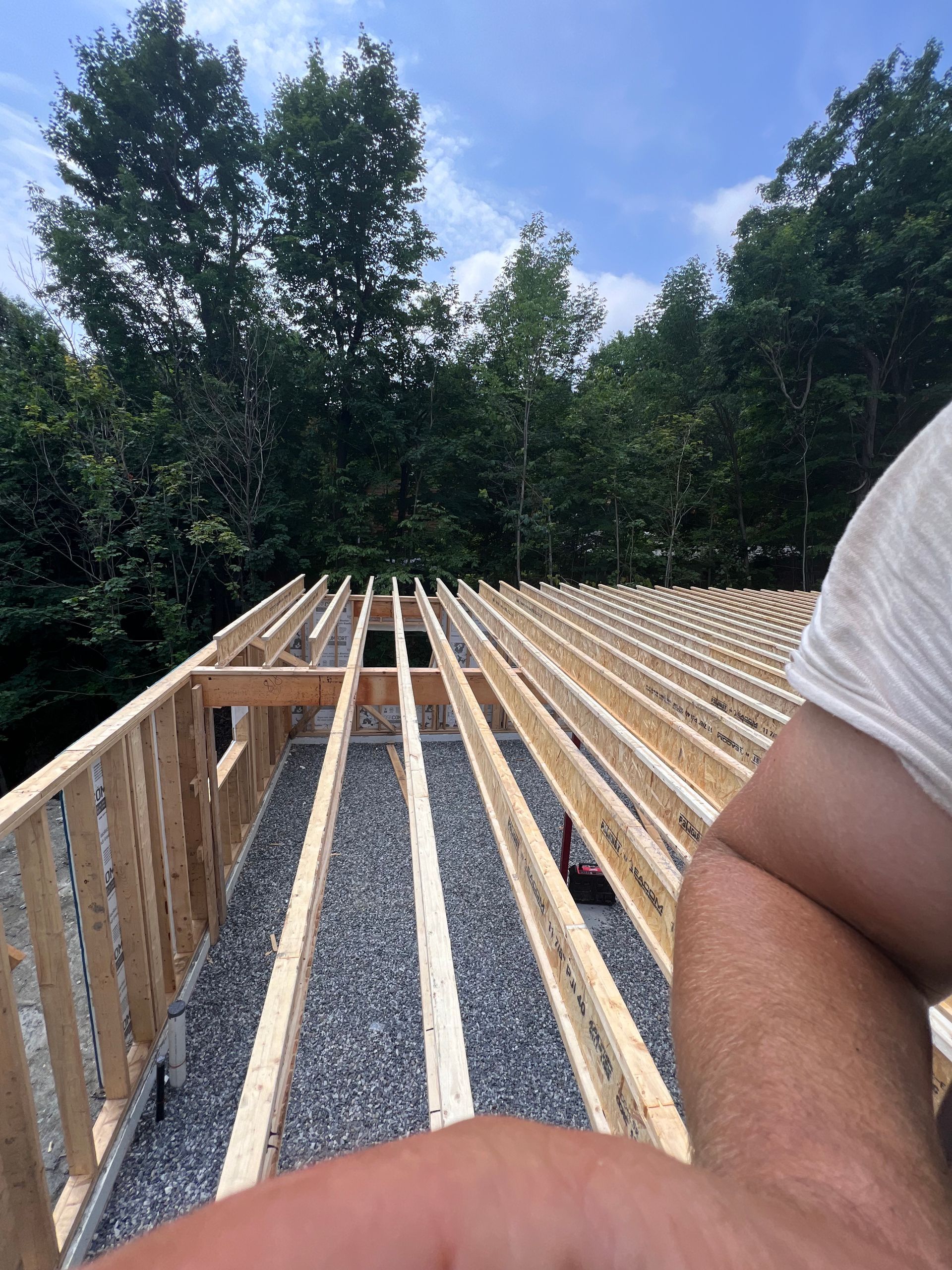 Charpente de toit en bois en construction à côté d'une forêt, avec du gravier en dessous et un bras humain au premier plan.