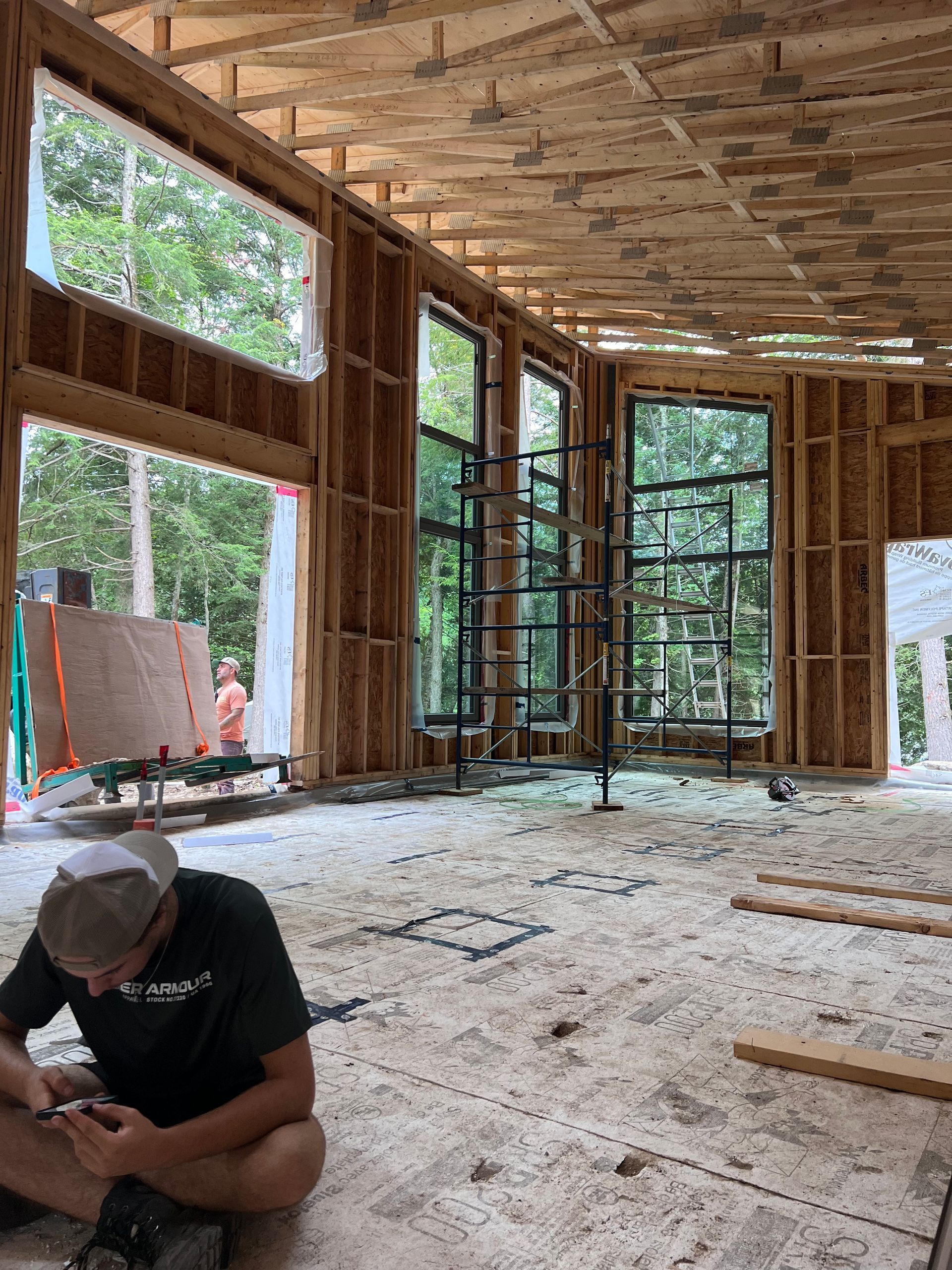 Intérieur d'une maison en construction, avec charpente apparente, fenêtres et un ouvrier accroupi sur le sol.