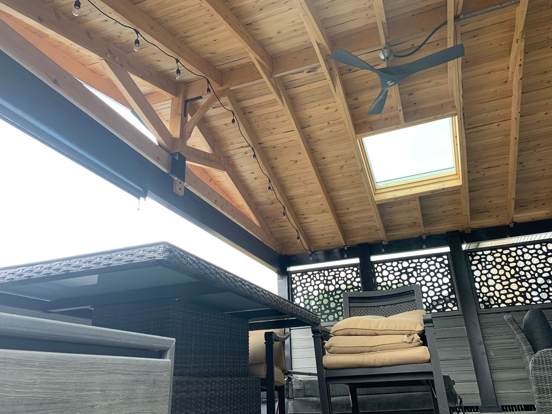 Toit de terrasse en bois avec puits de lumière et ventilateur de plafond au-dessus de meubles en osier et de coussins empilés.