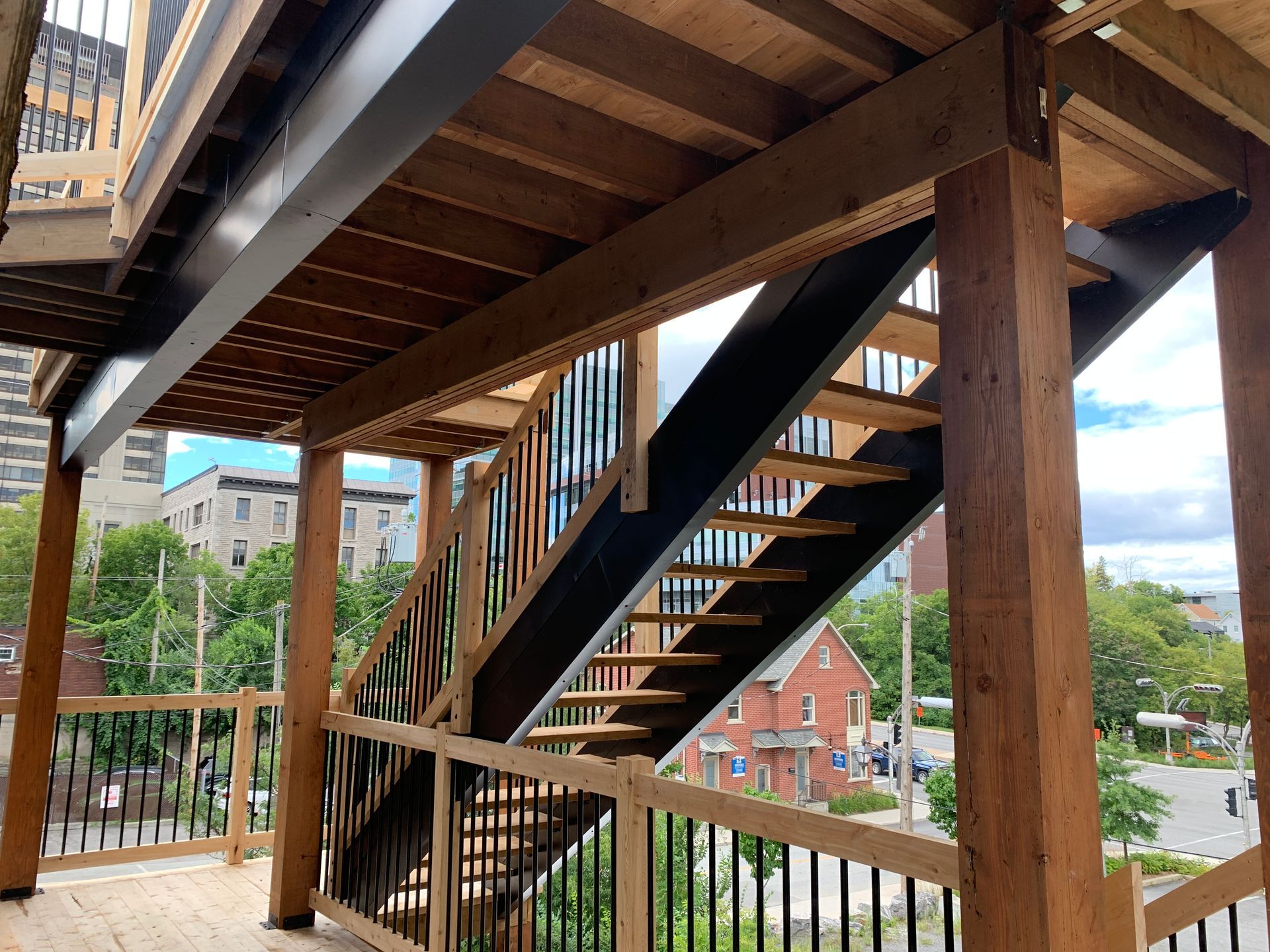 Escalier extérieur en bois sous une terrasse, donnant sur une ville aux bâtiments en briques et aux arbres.