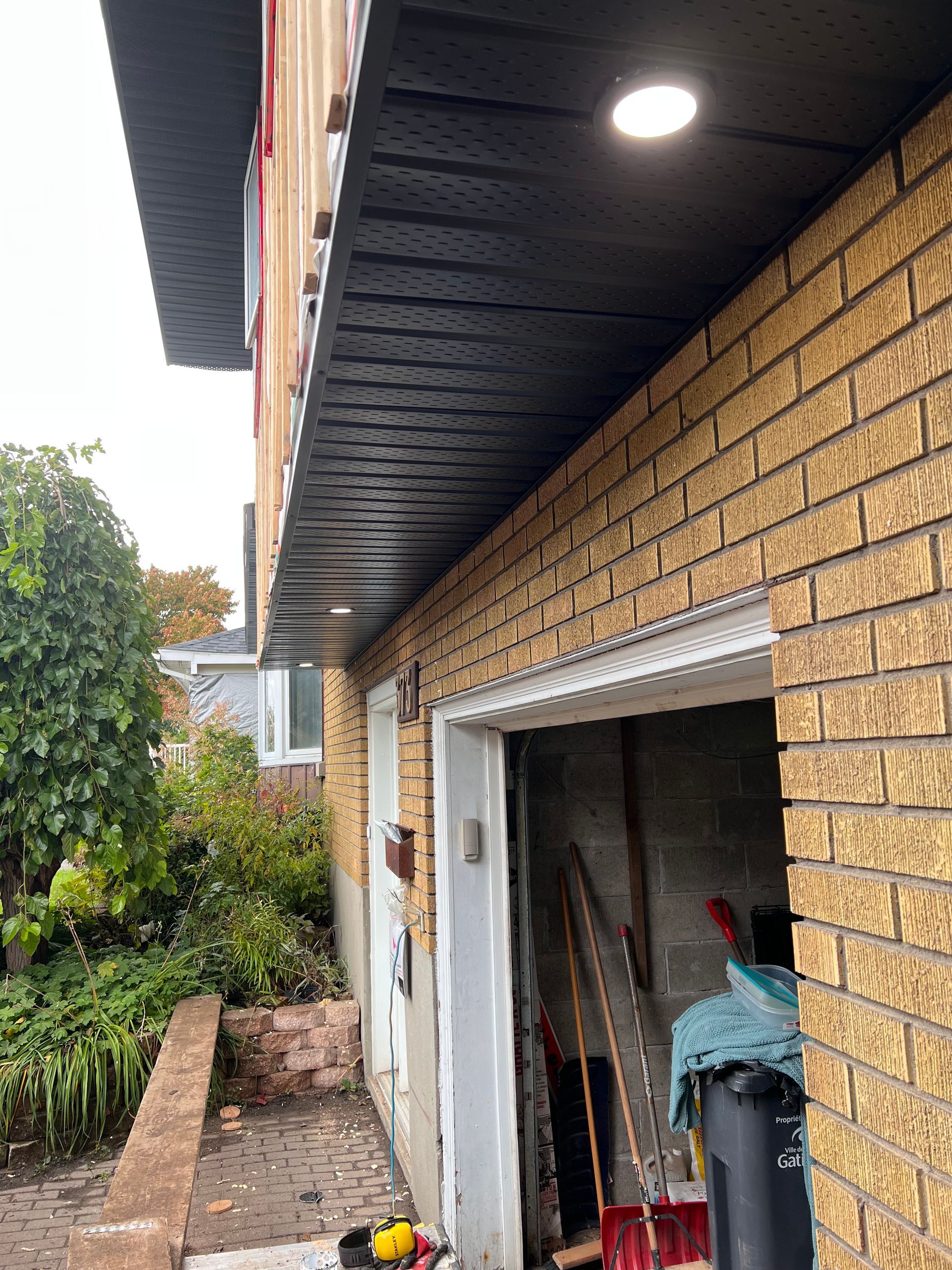 Vue latérale d'une maison en briques avec une porte de garage ouverte, des avant-toits sombres et une lumière allumée sous l'avancée de toit.