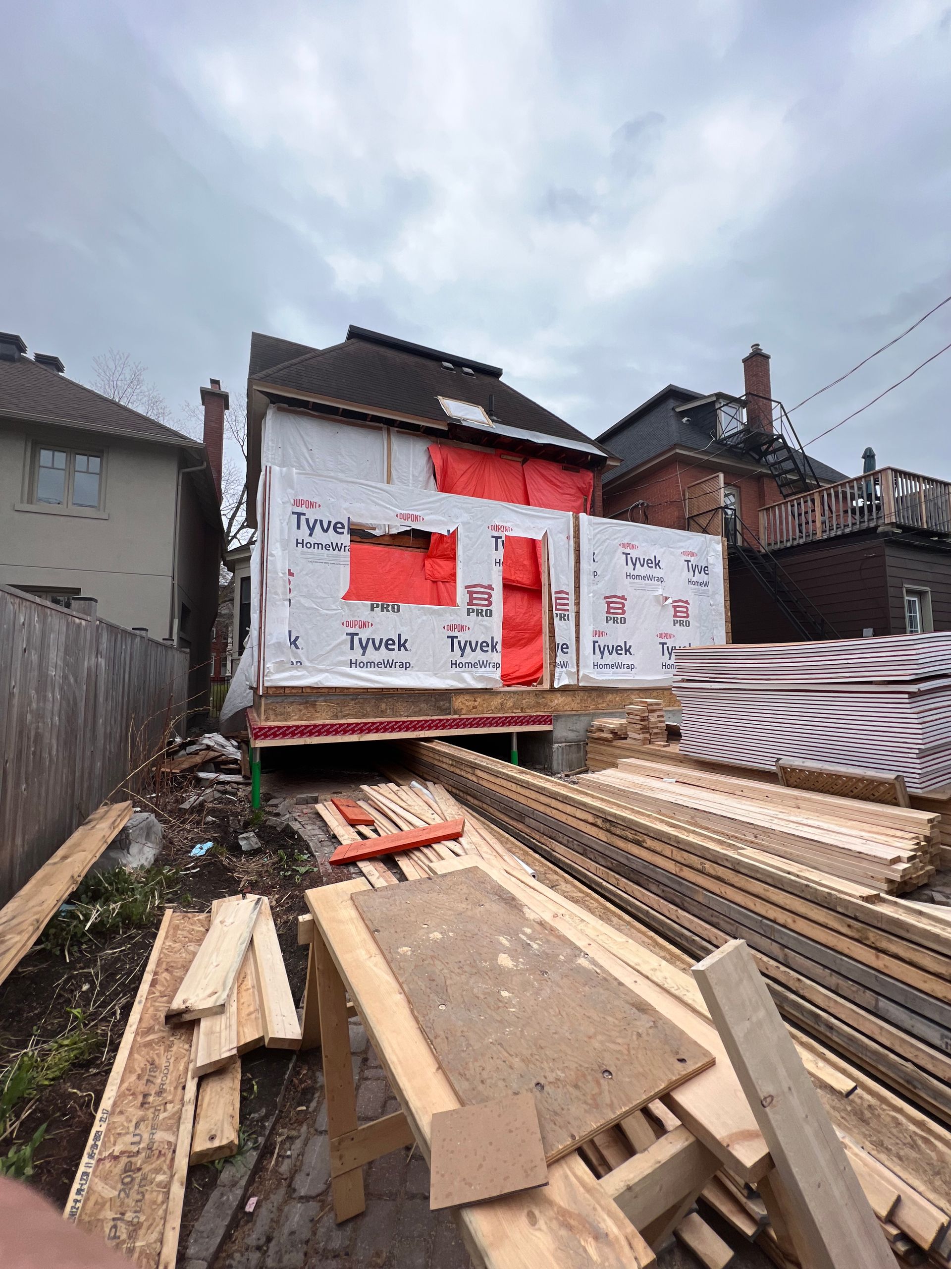 Chantier de construction avec une maison partiellement construite, charpente apparente et bois empilé au premier plan.