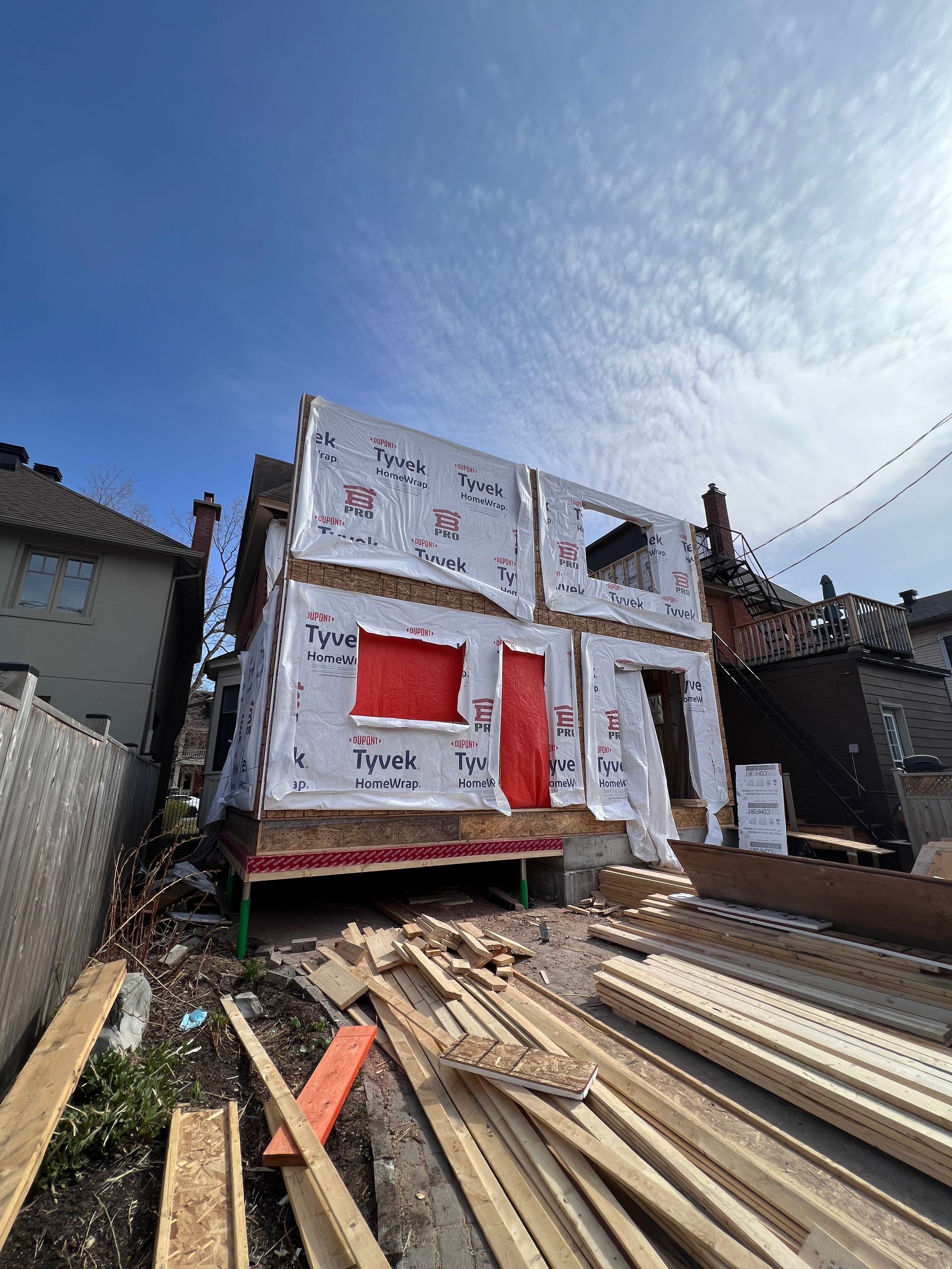 Maison partiellement construite avec un revêtement blanc, des rideaux rouges et du bois empilé au premier plan sous un ciel bleu