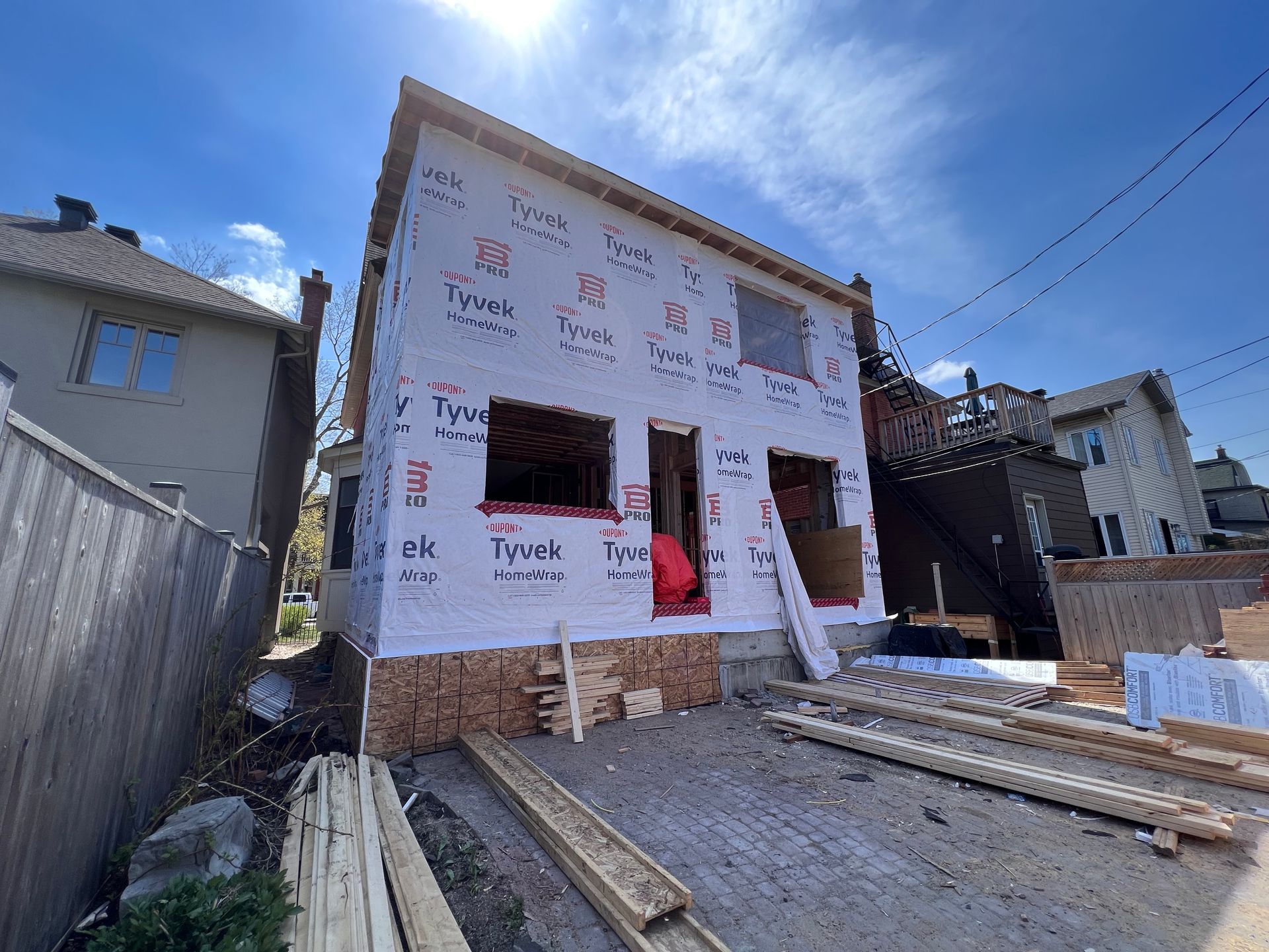 Maison partiellement construite, recouverte d'un pare-vapeur Tyvek, avec une ossature en bois et des matériaux de construction visibles à l'avant.