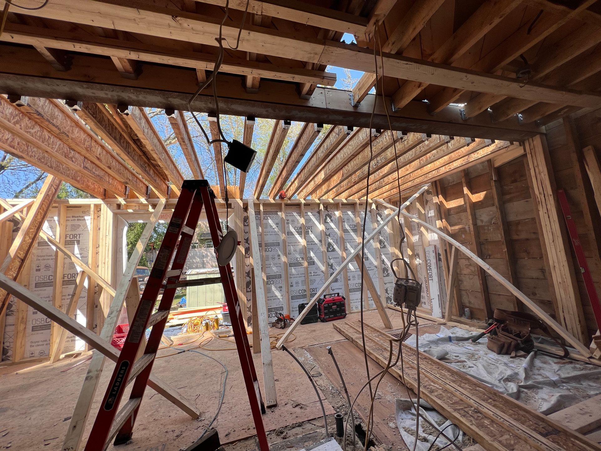 Intérieur d'une maison en construction avec une charpente en bois apparente, une échelle rouge et des débris au sol.