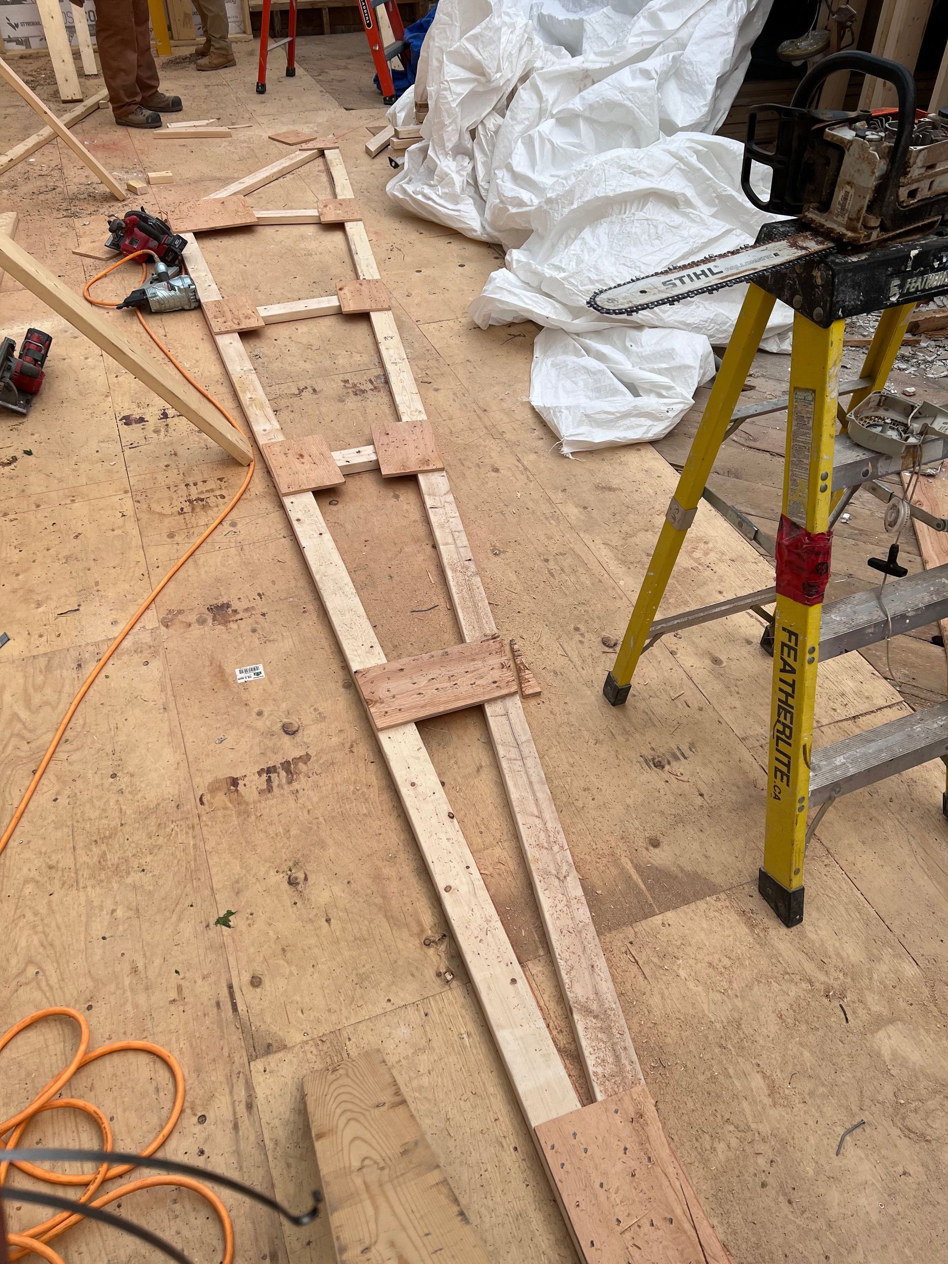 Zone de construction avec des étais en bois au sol, un escabeau jaune et une rallonge électrique orange enroulée.