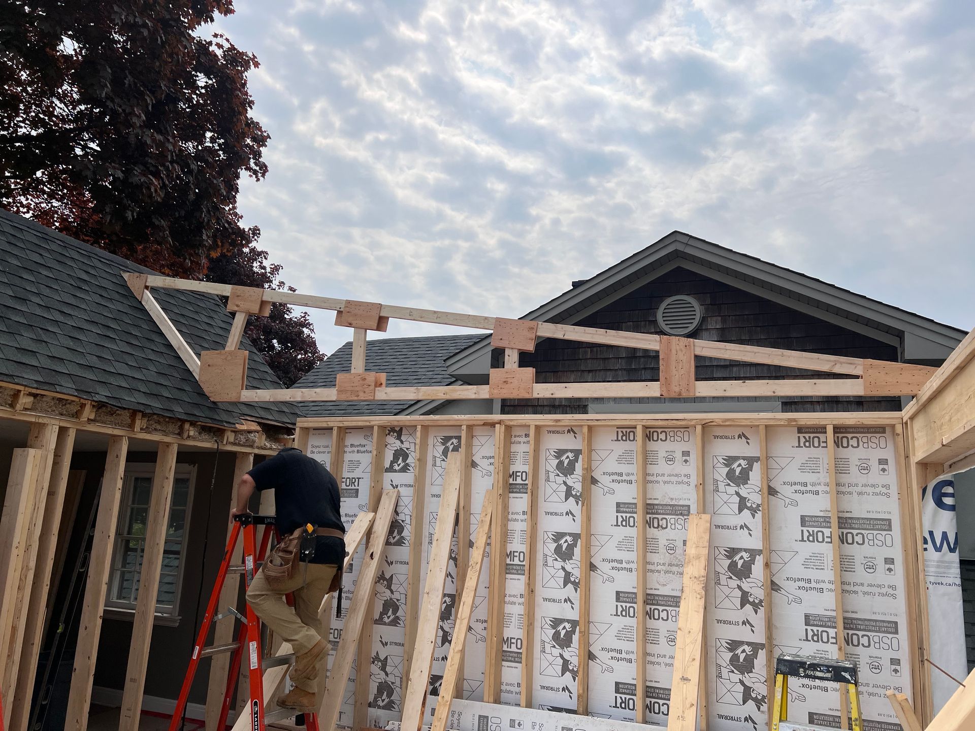 Un ouvrier sur une échelle travaille à la charpente d'un toit de maison, avec l'isolation des murs apparente et des murs partiellement construits.
