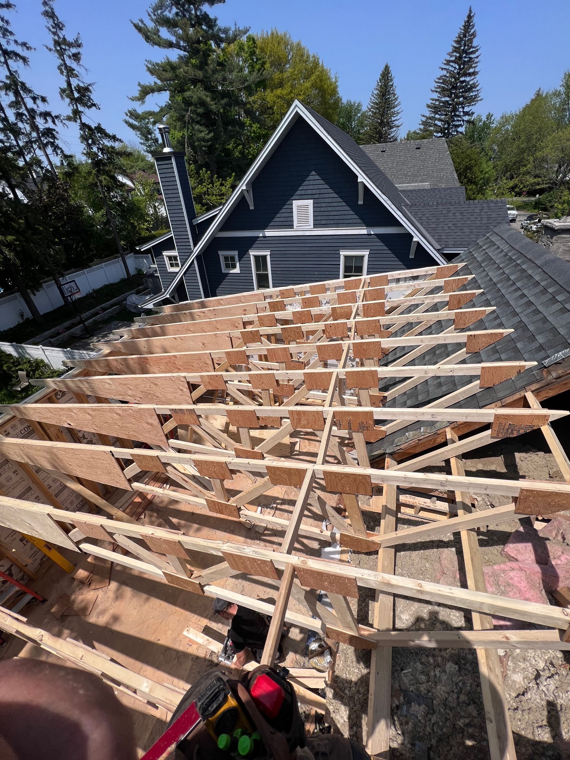 Toiture en construction avec charpente apparente à côté d'une maison bleu foncé et d'arbres