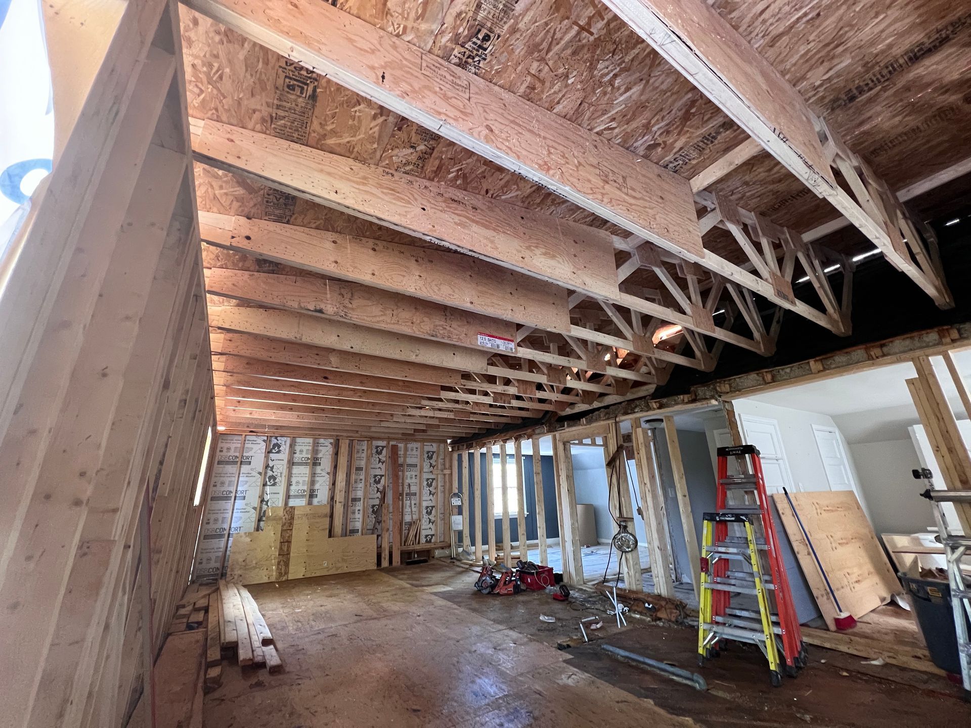 Pièce inachevée avec poutres apparentes au plafond, échelles, outils et matériaux de construction au sol.