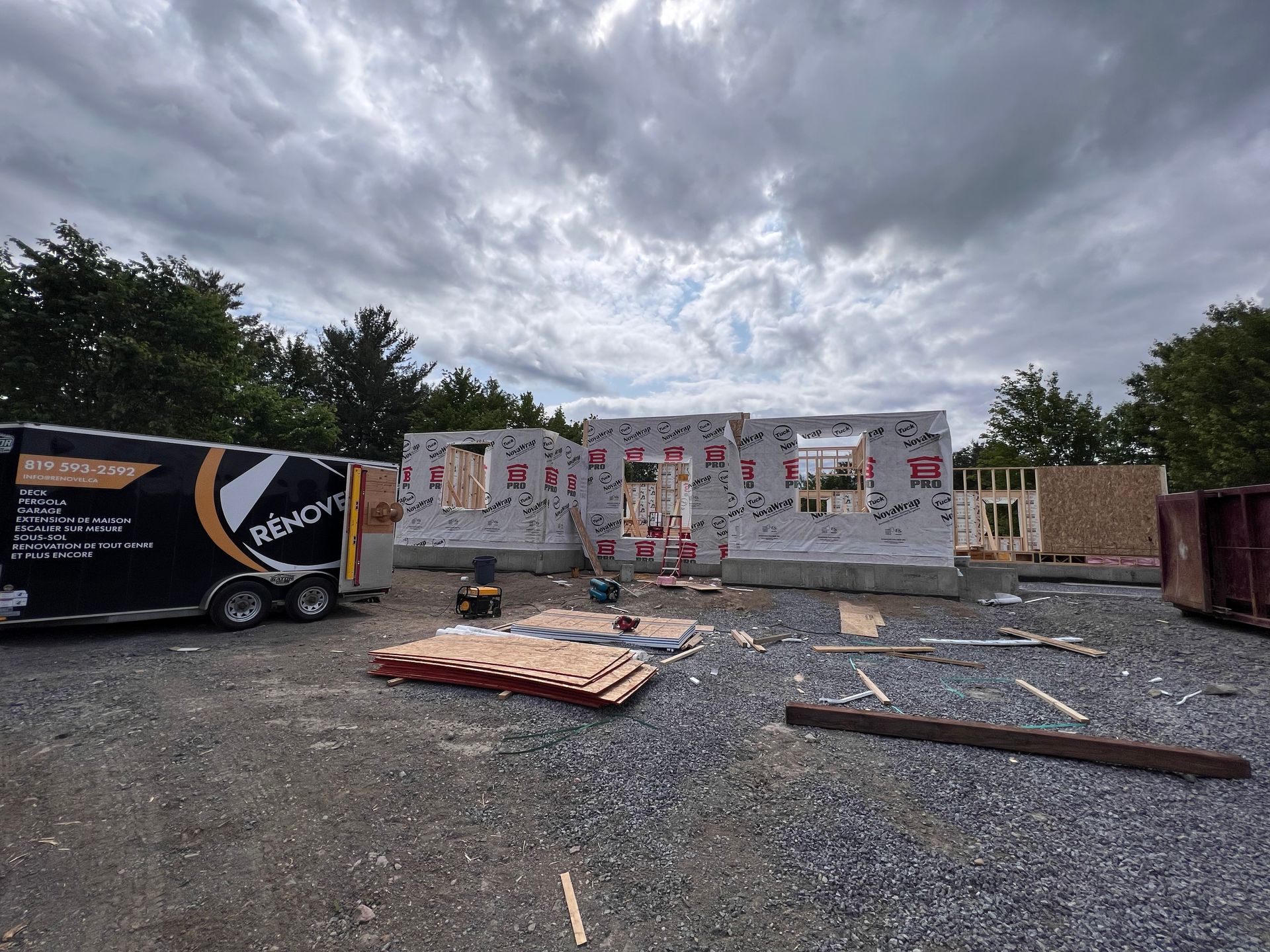 Chantier de construction endommagé par la tempête : débris, remorque et charpente apparente sous un ciel nuageux.