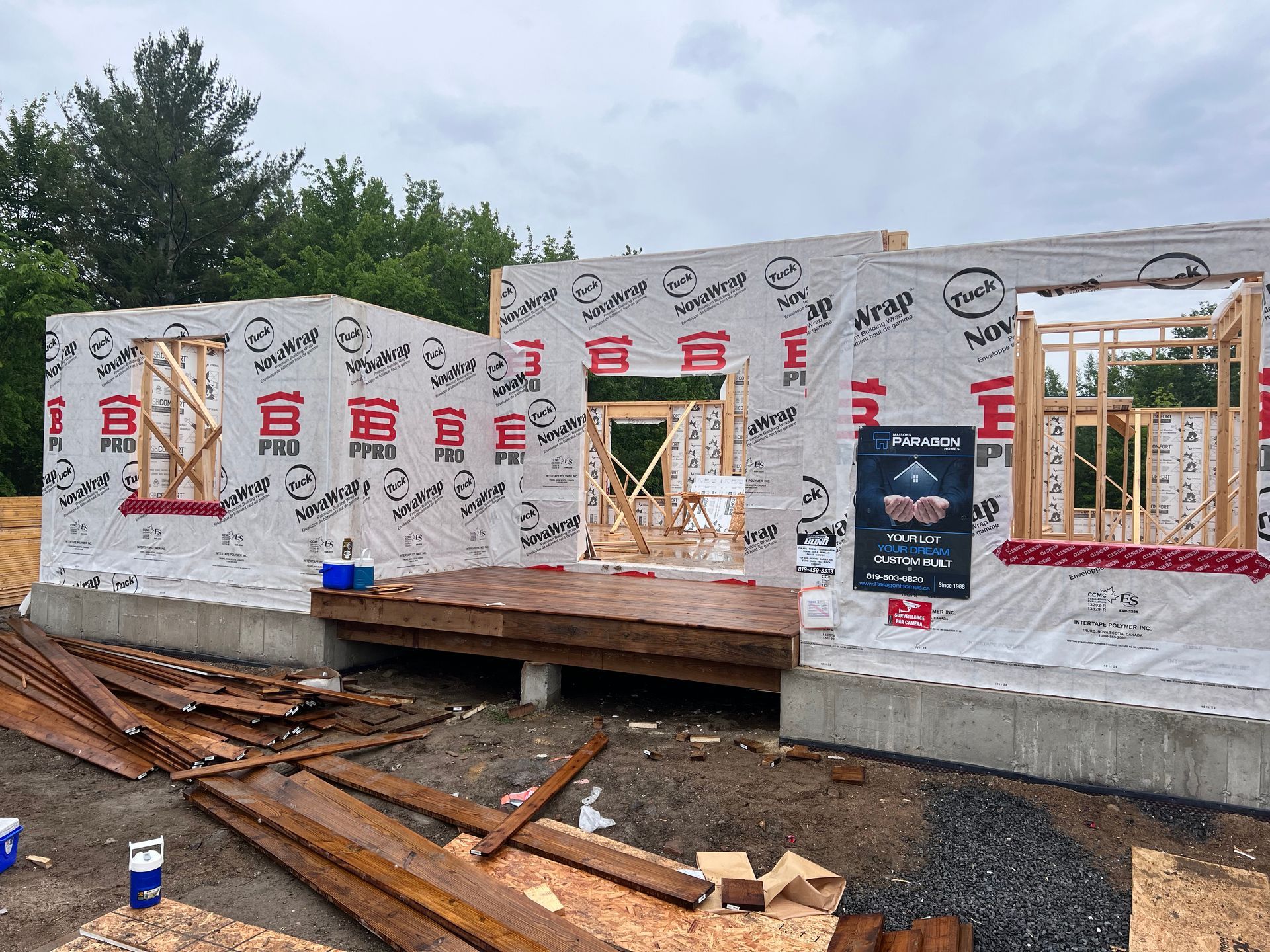 Maison en construction avec murs extérieurs recouverts de bâches, ouvertures pour fenêtres et bois empilé au sol.