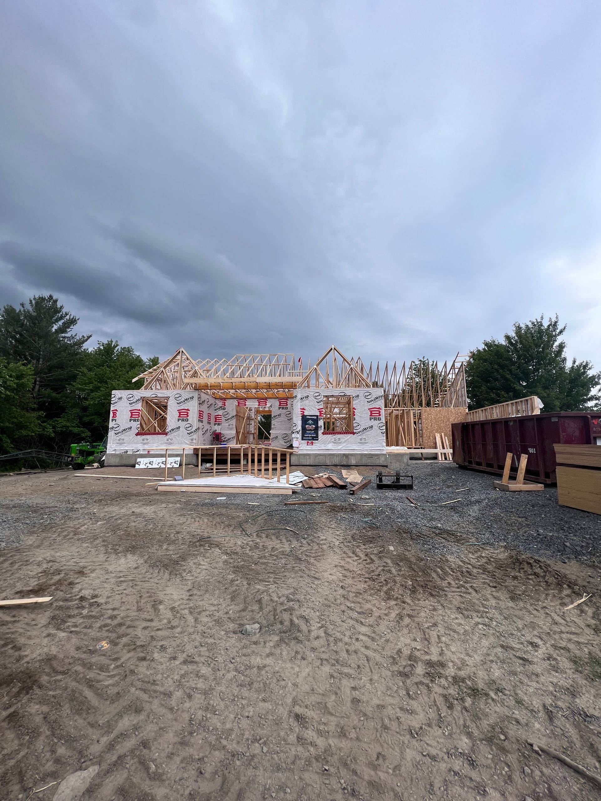 Ossature de maison partiellement construite sur un terrain gravillonné sous un ciel nuageux, avec des matériaux éparpillés autour