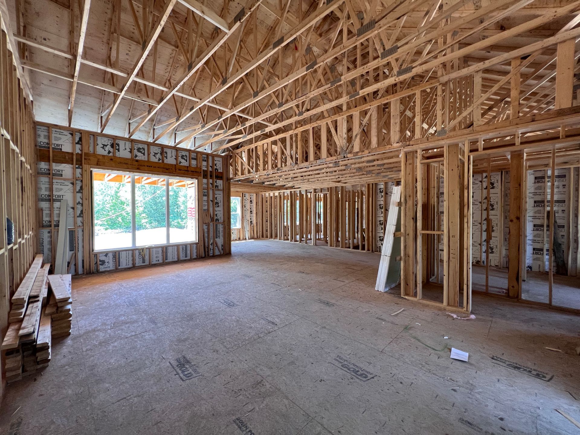 Intérieur d'une maison inachevée avec charpente en bois apparente, fermes de plafond ouvertes et sol en béton brut.