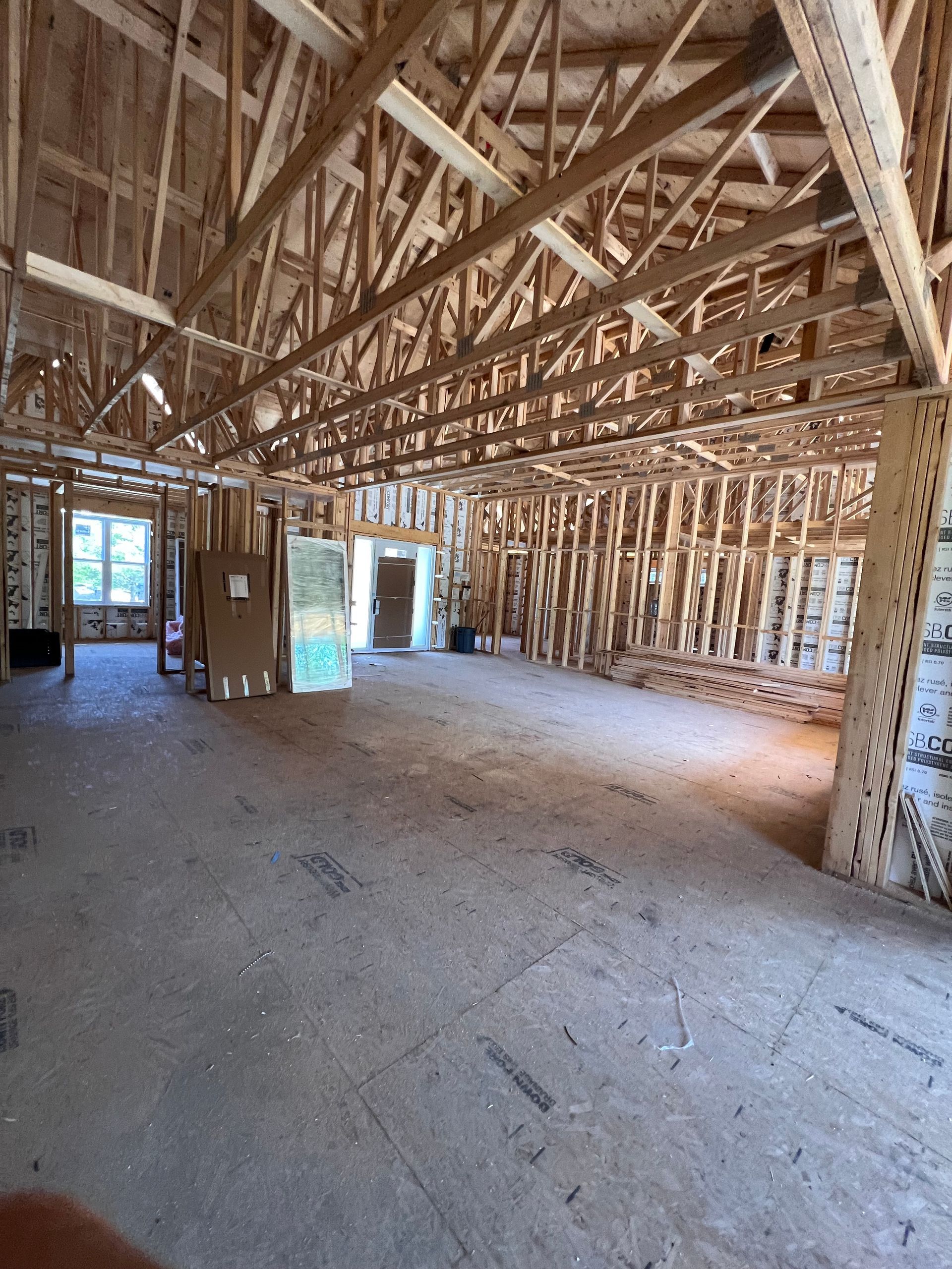 Intérieur d'une maison en bois inachevé, avec charpente apparente, fermes et sol en béton près d'une porte et de fenêtres.