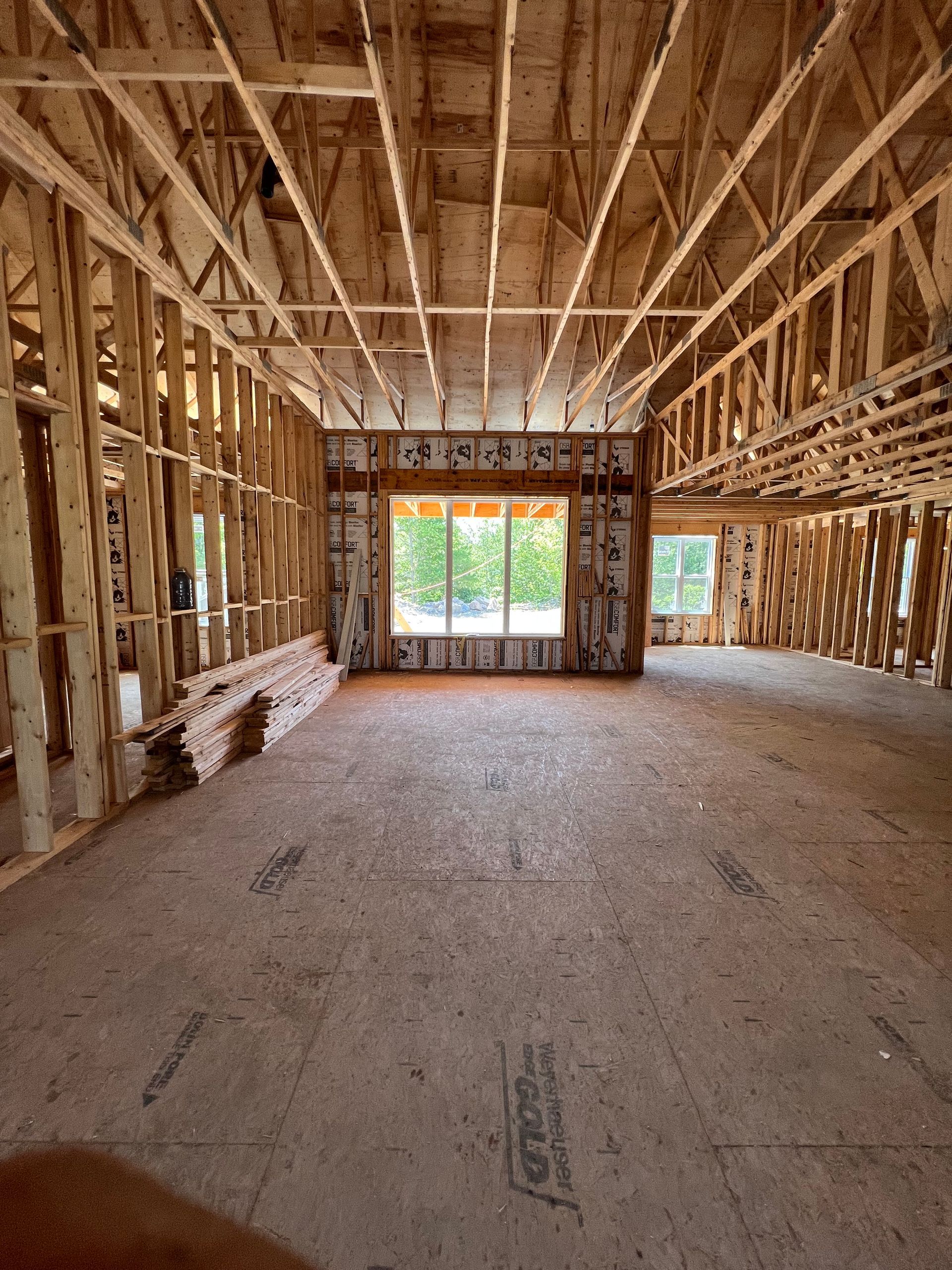 Intérieur d'une maison inachevée, avec une charpente en bois apparente et une grande fenêtre au fond.