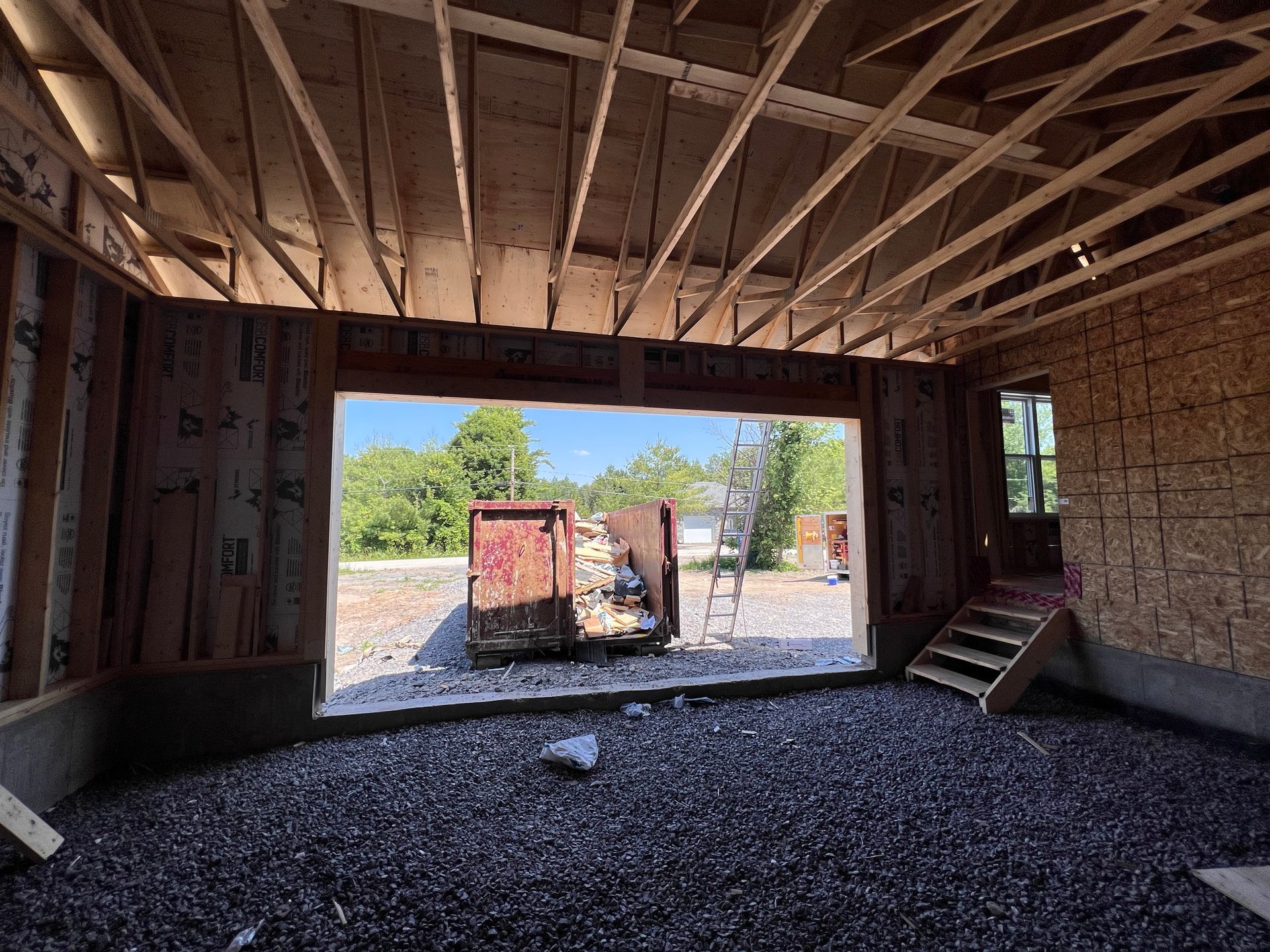 Intérieur d'un bâtiment inachevé avec charpente apparente, sol en gravier et une grande porte ouverte donnant sur un chantier.