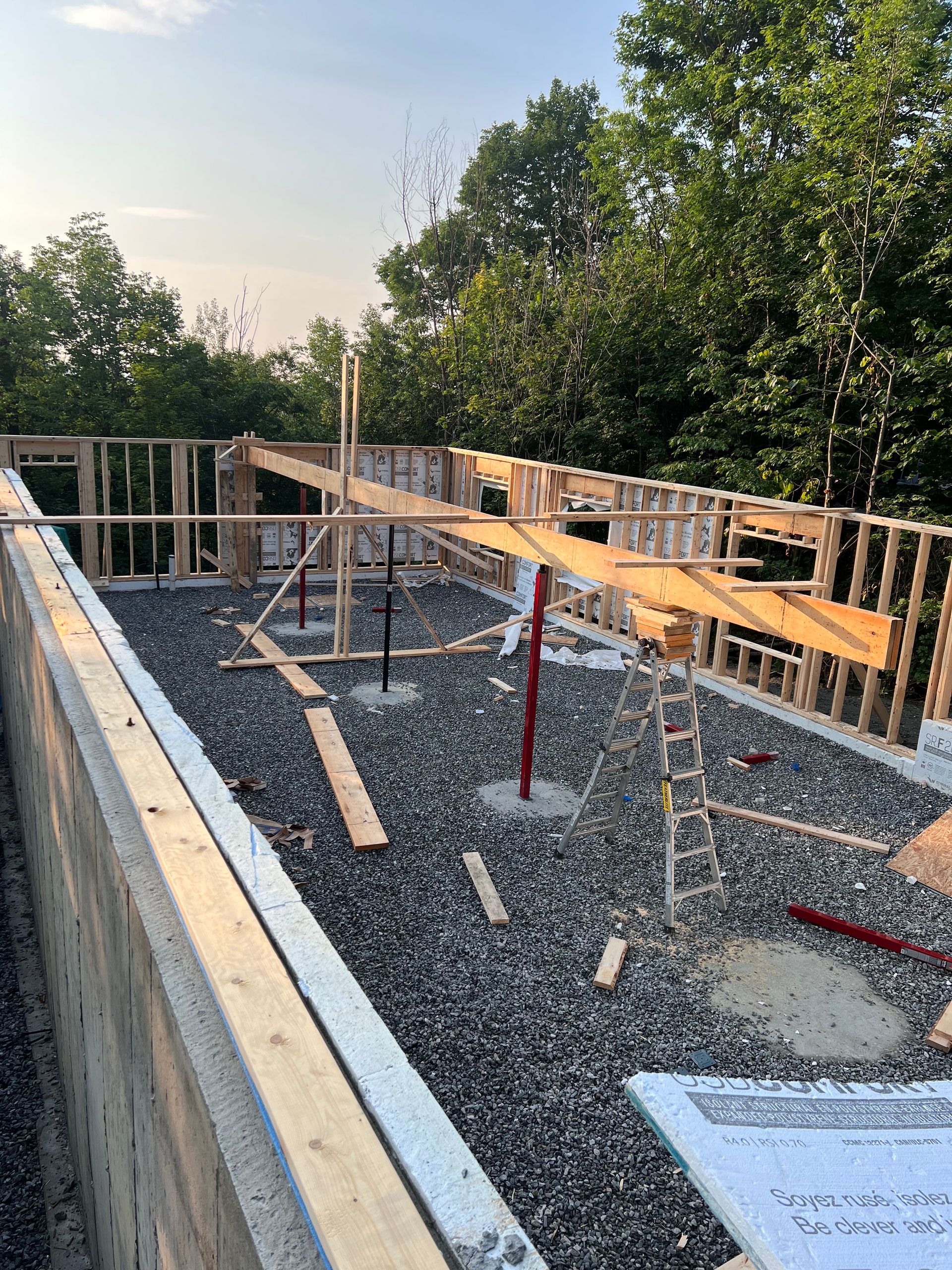 Charpente de toit en construction avec poutres en bois, échafaudages et gravier sur une structure partiellement construite au coucher du soleil