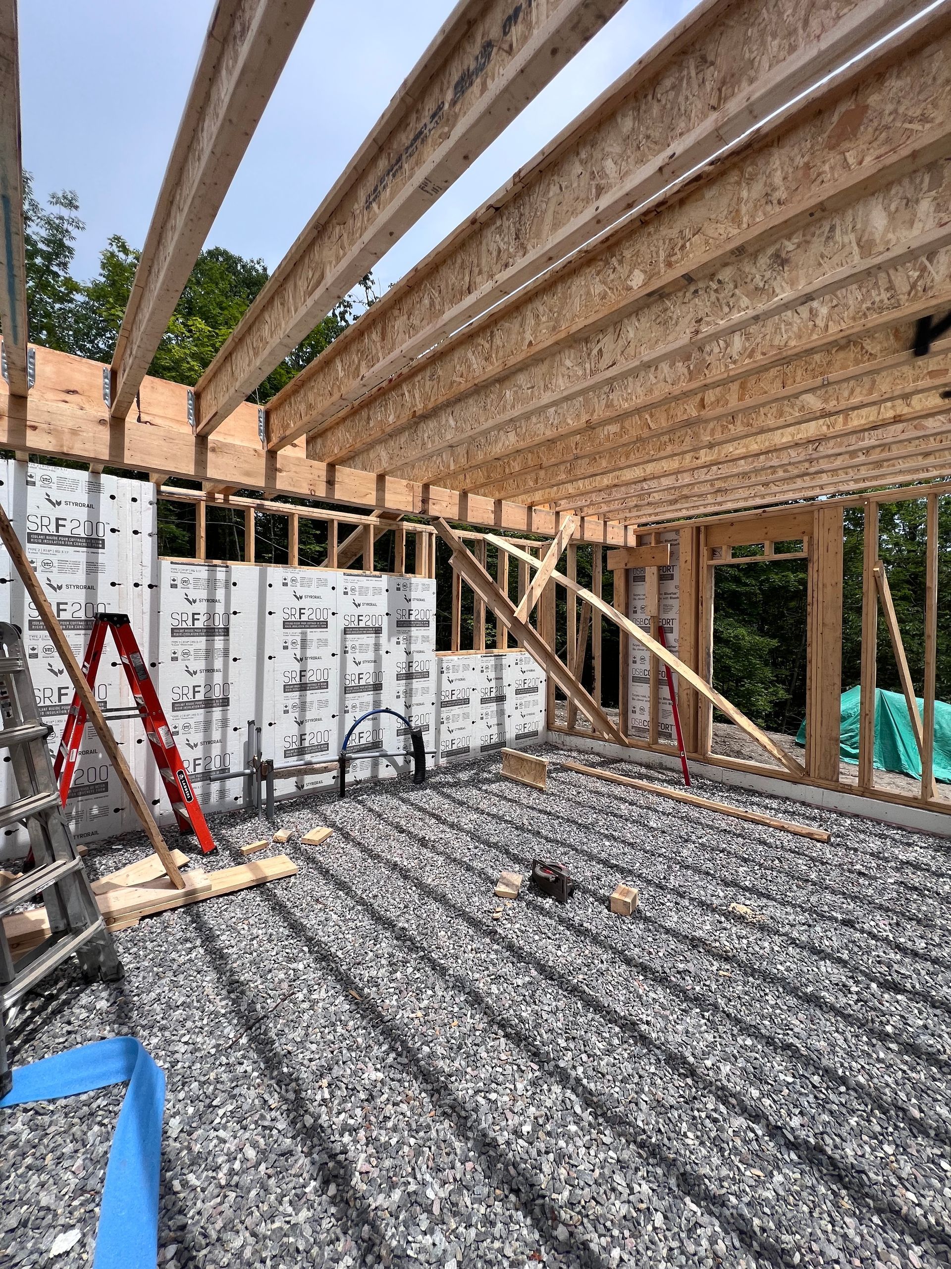 Chantier de construction avec charpente apparente, panneaux muraux blancs et échelle rouge sur un sol en gravier