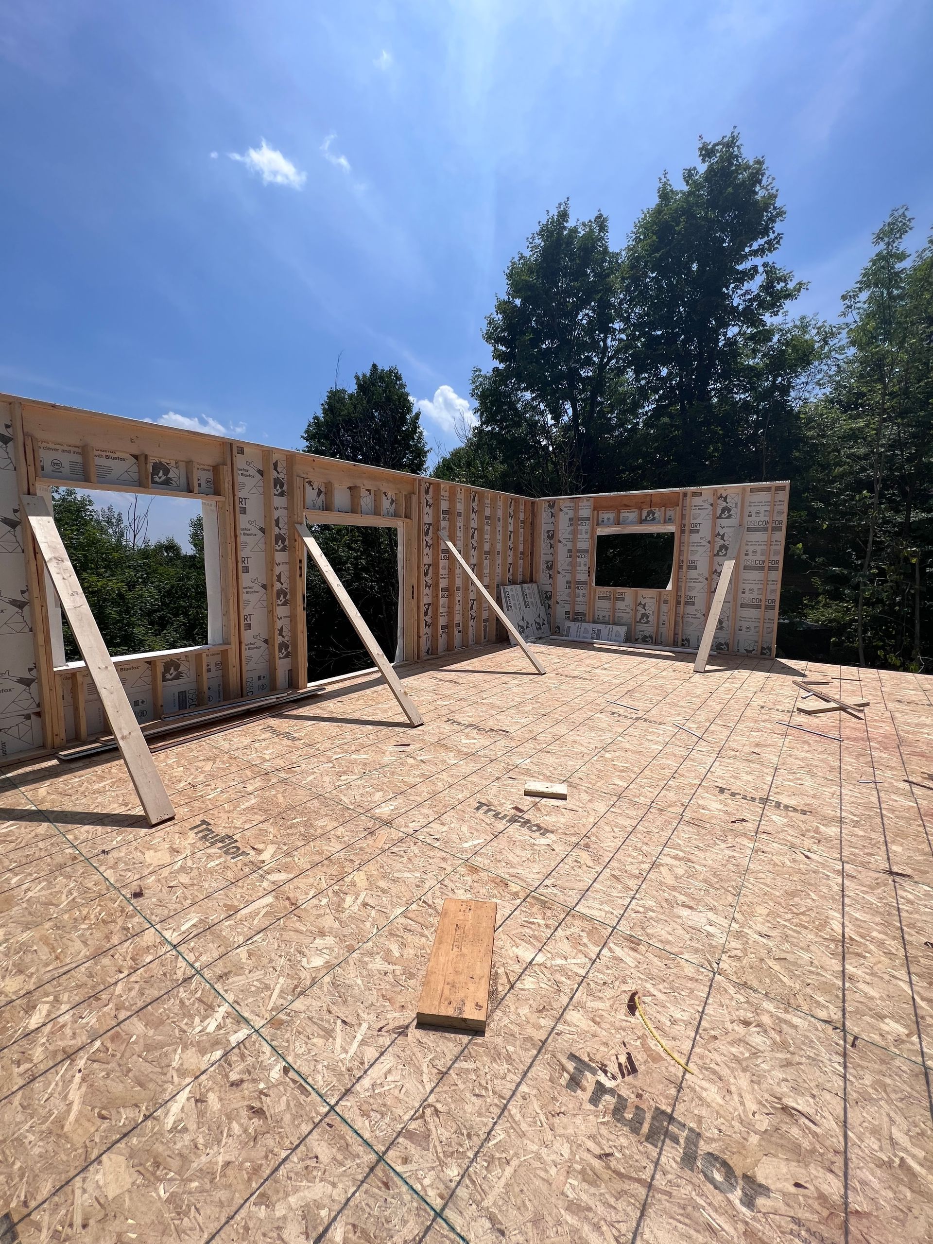 Intérieur d'une maison inachevée avec charpente apparente, plancher en contreplaqué et ouvertures pour les fenêtres et les portes sous un ciel bleu