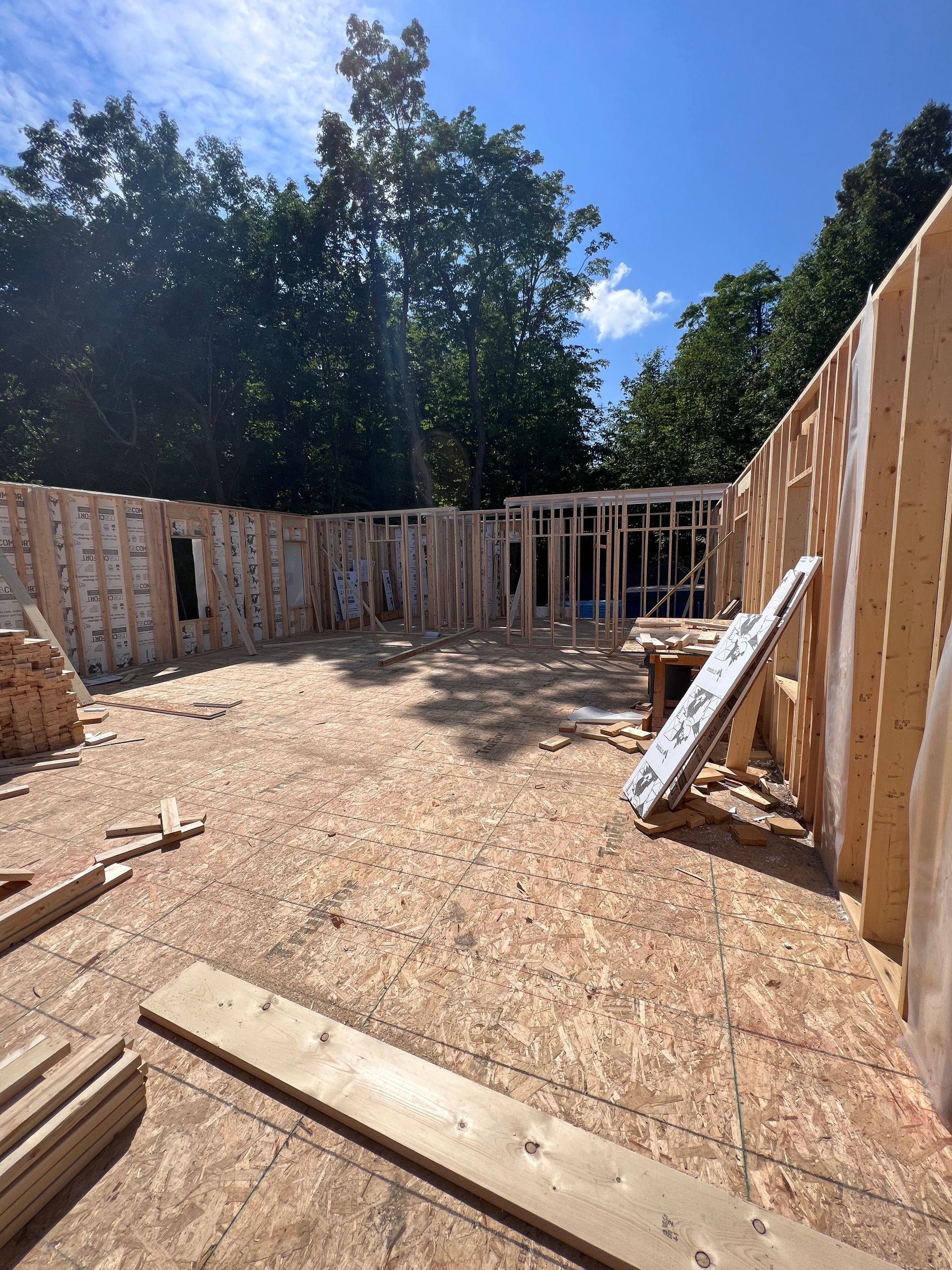 Intérieur d'une maison partiellement construite, avec charpente apparente et plancher en contreplaqué, sous un ciel bleu.